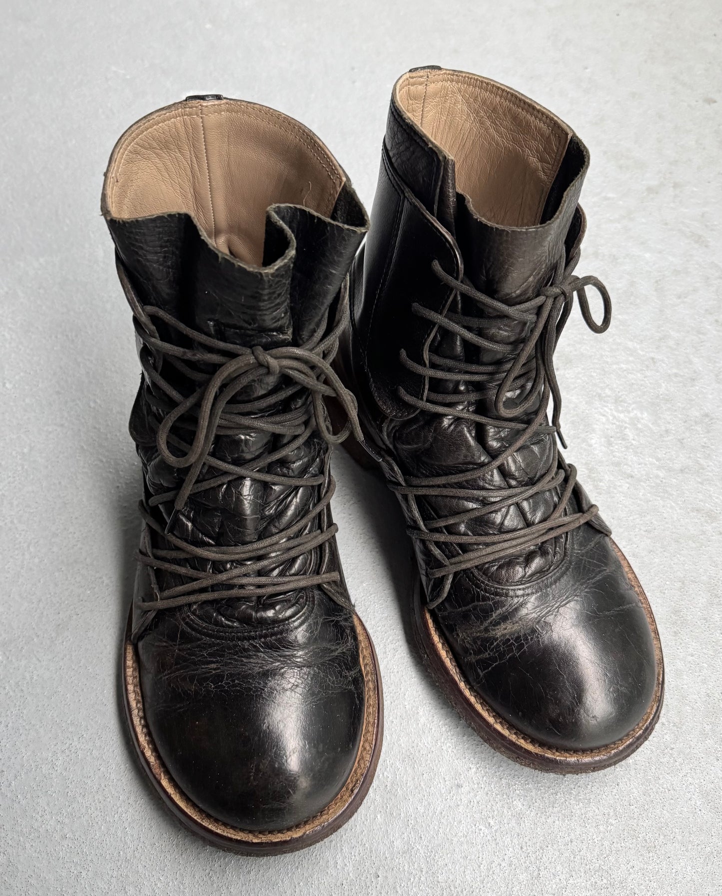 A pair of worn Mihara Yasuhiro Early 00s Multi-laced Crepe Sole Boots, showcasing distinctive texture and visible creases, rests on a light gray surface. They embody avant-garde fashion with subtle deconstructed elements as they angle slightly towards each other.