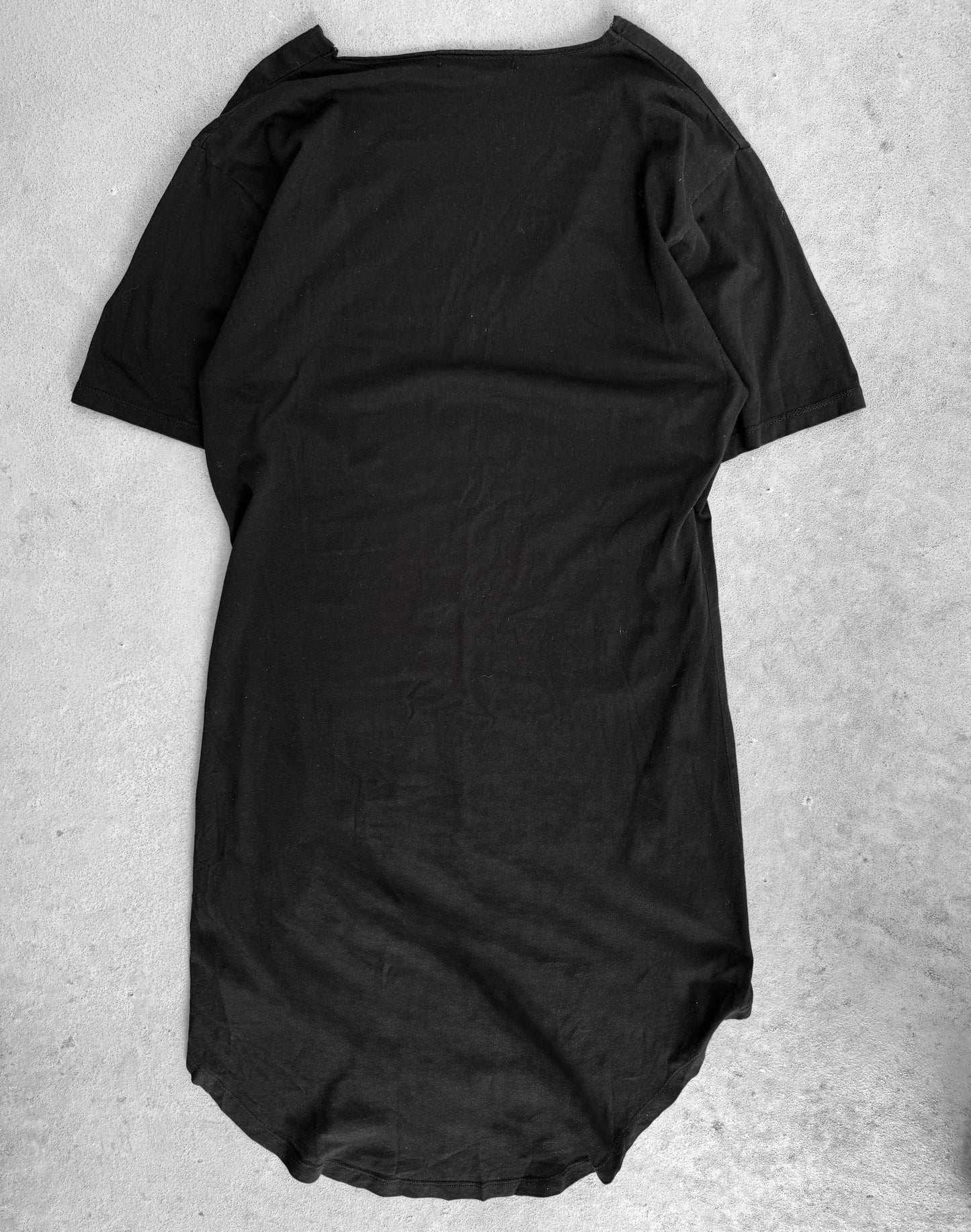 The CIVARIZE Early 00s Floral Skull Graphic Long Curve T-shirt in black rests on a light gray surface. It showcases visual kei-inspired designs, a round neckline, and a slightly curved hem, with the thin fabric revealing the background texture for an alternative fashion feel.