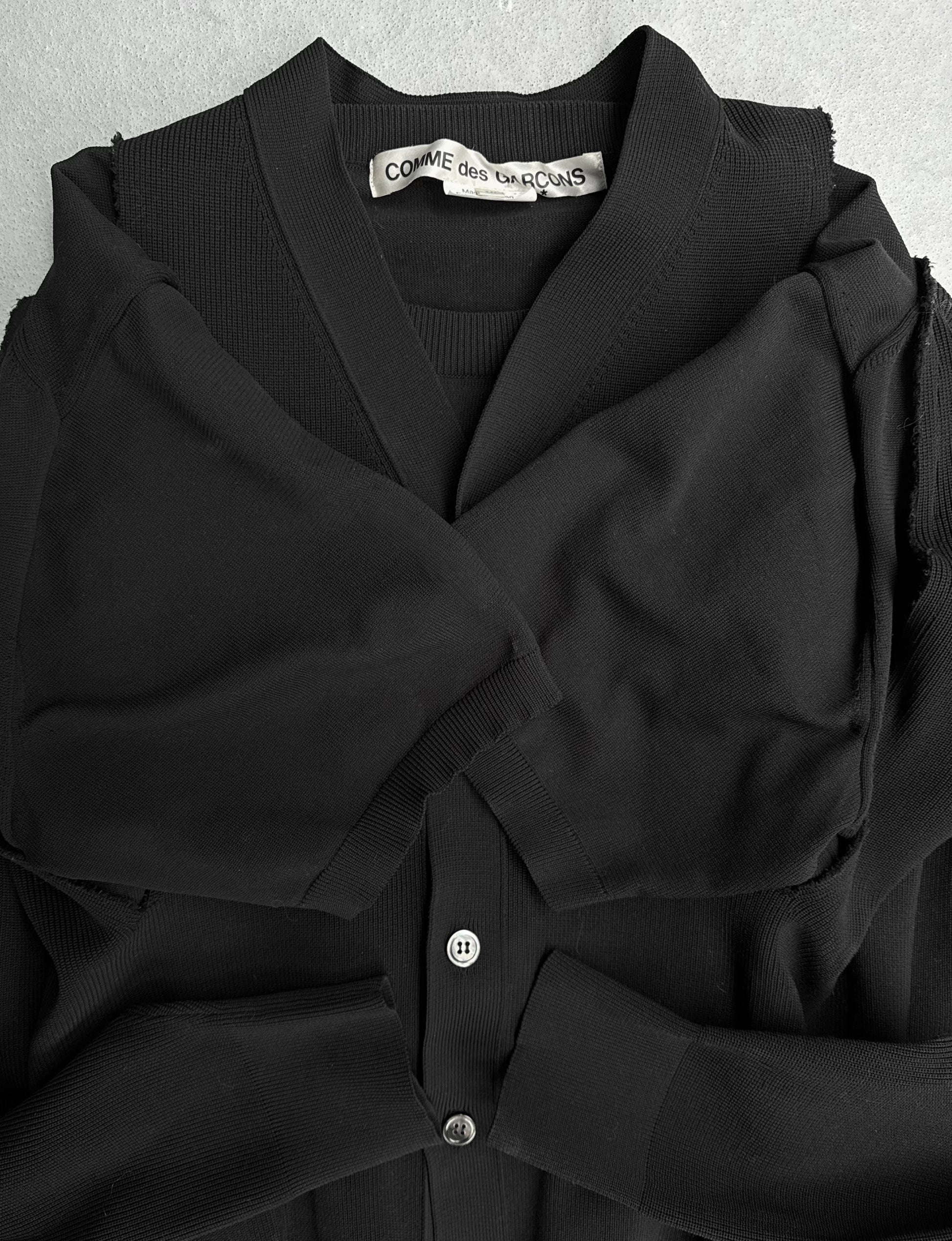 The COMME des GARÇONS SS19 Deconstructed 2-in-1 Cardigan, in classic black, embodies avant-garde fashion with its signature label at the neckline, neatly folded on a gray surface—a testament to its exclusive Dover Street Market availability.