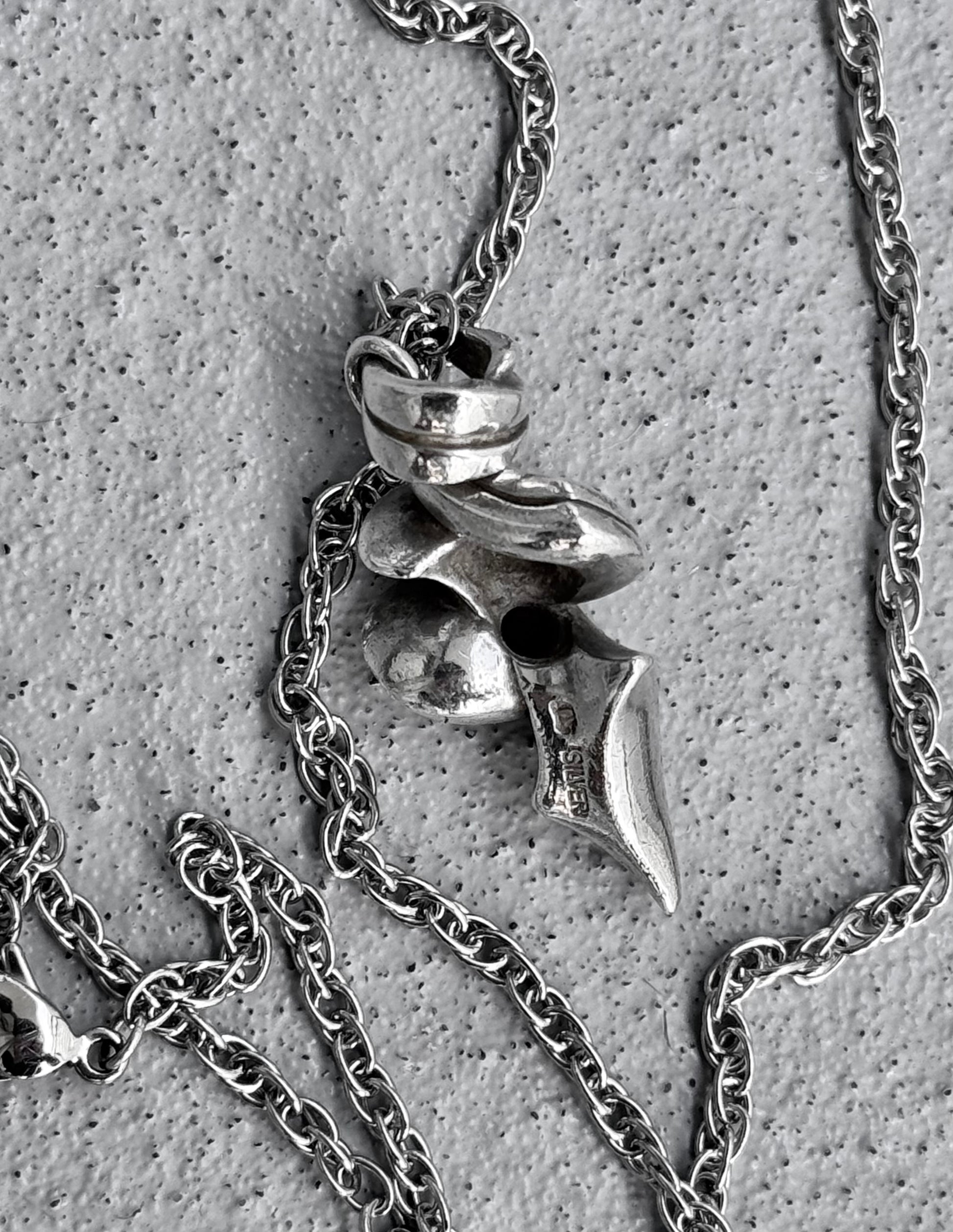 The M’s Collection (BLESS) Early 00s Silver Tribal Twist Pendant Chain showcases BLESSs flair for conceptual fashion, with abstract shapes hanging on a silver chain. Set against a textured grey surface, it combines smooth and pointed elements to enhance its intricate and artistic design.