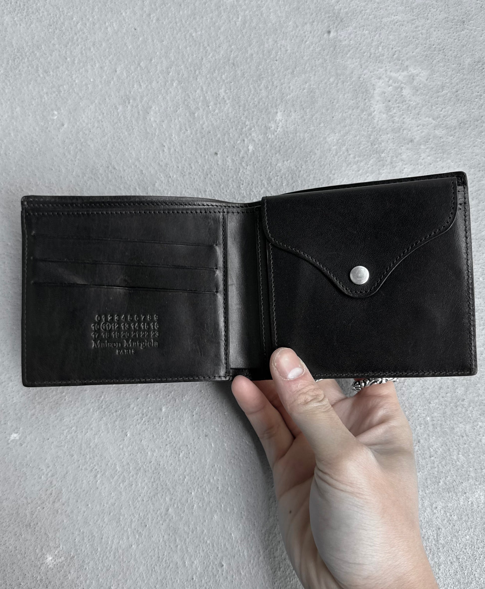 A hand displays a Maison Martin Margiela AW15 Distressed Calfskin Leather Bi-fold Wallet (OneSize, black, 8/10 condition) with snap-button coin pouch and card slots against a textured gray background.