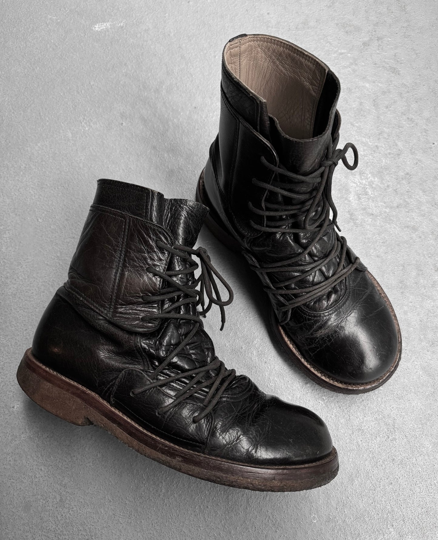 MiharaYasuhiro Early 00s Multi-laced Crepe Sole Boots, with their worn black leather and brown soles, sit laced on a gray concrete background. These boots flaunt deconstructed features and creased leather, embodying an avant-garde aesthetic through their rugged design.