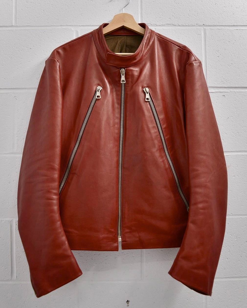 The Maison Martin Margiela AW00 Red Cowhide Leather 5-zip Rider Jacket, with two slanted front zippers and a simple collar, hangs on a wooden hanger against a white textured wall, showcasing the brands avant-garde yet understated design.