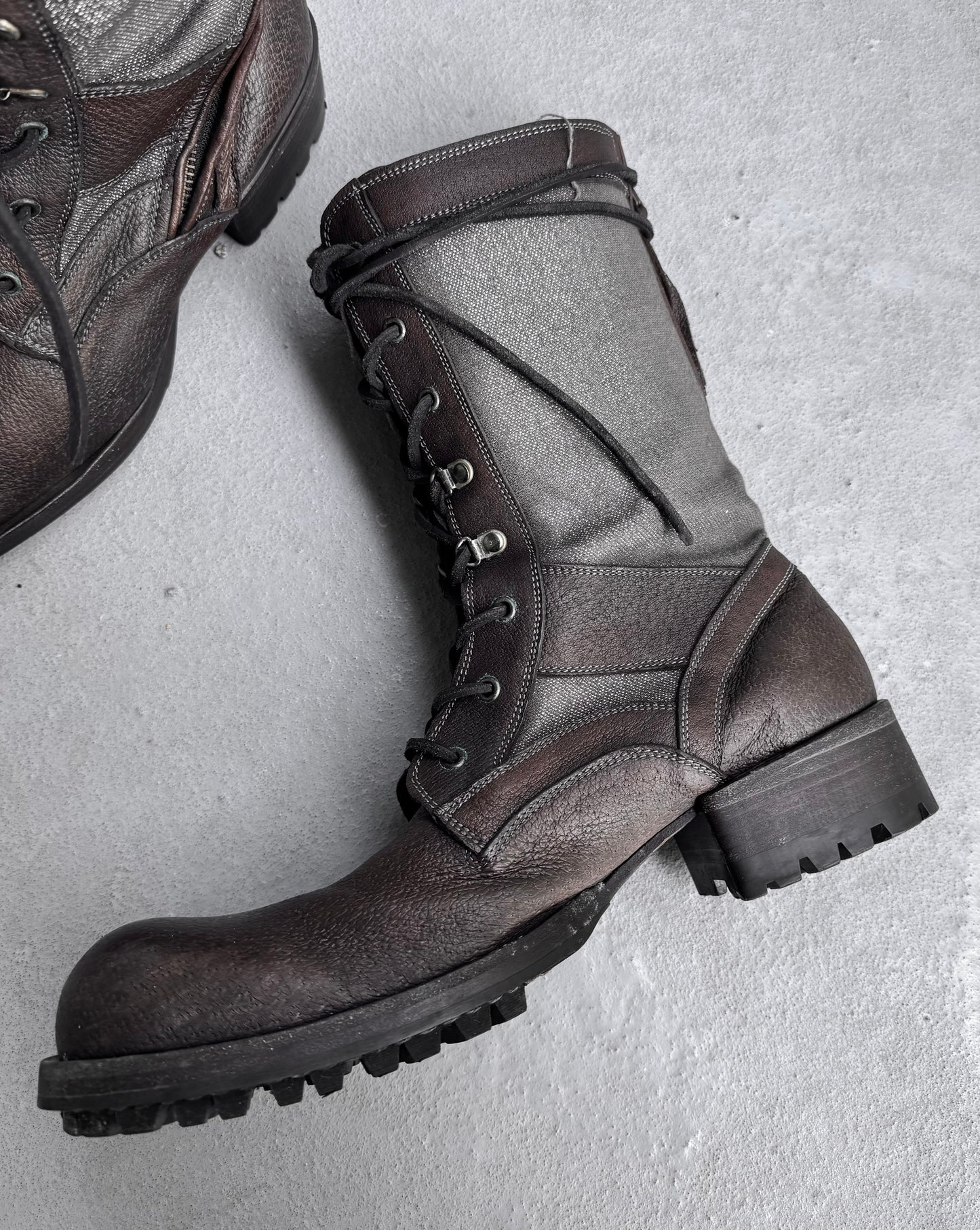 A pair of Kyoji Maruyama Early 00s Brown Tornado Zip Combat Boots with thick rubber soles and metal eyelets lie on a concrete floor, embodying Japanese fashion. One boot is fully visible with laced-up details; the other is partially visible in the top left corner.