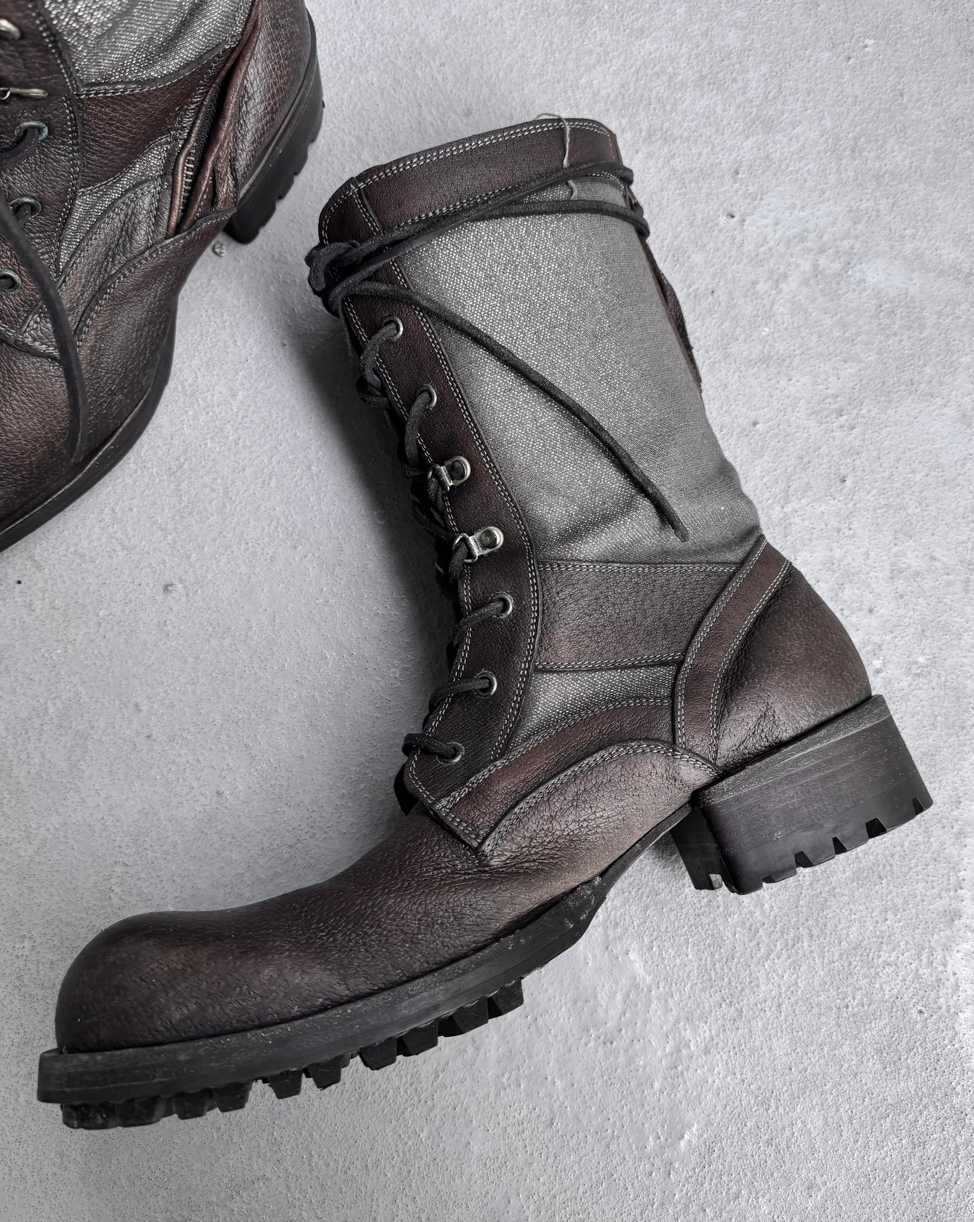 A pair of Kyoji Maruyama Early 00s Brown Tornado Zip Combat Boots with thick rubber soles and metal eyelets lie on a concrete floor, embodying Japanese fashion. One boot is fully visible with laced-up details; the other is partially visible in the top left corner.