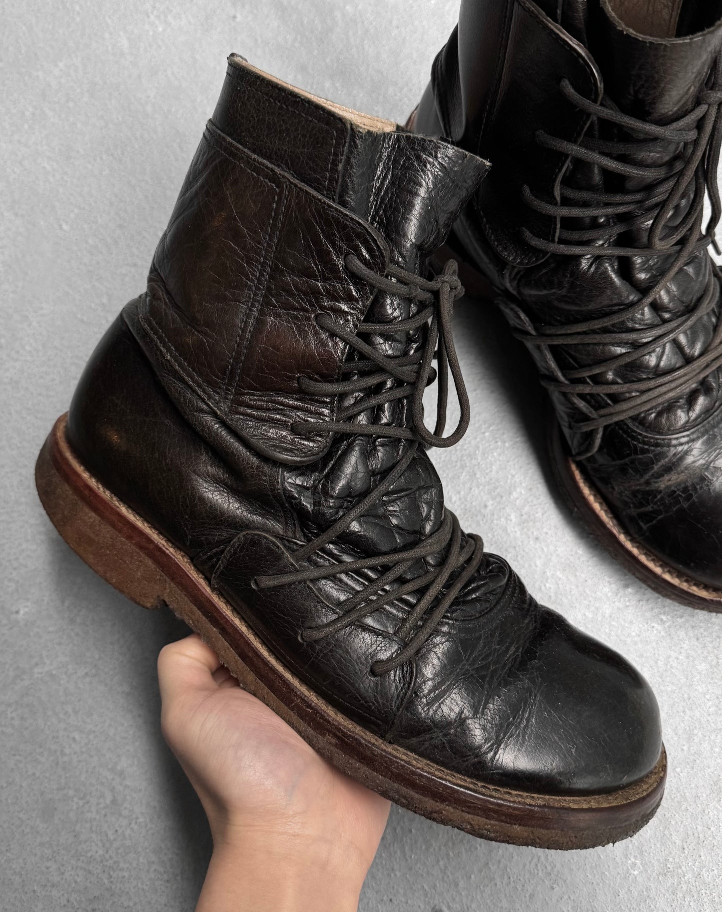 A hand holds a black, worn Mihara Yasuhiro Early 00s Multi-laced Crepe Sole Boot with brown soles against a plain backdrop, highlighting its deconstructed style. Another boot is partially visible, evoking avant-garde fashion sophistication.