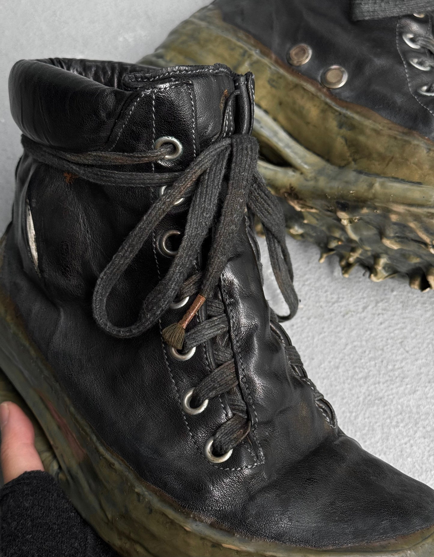 A close-up of a worn DRIEW Carol Christian Poell ‘AM/2685P ROOMS-PTC/12’ object-dyed kangaroo leather high-top sneaker, size 6, with thick black laces, visible scuffs, and a distressed titanium prosthetic U-sole. Another similar sneaker is partially visible.