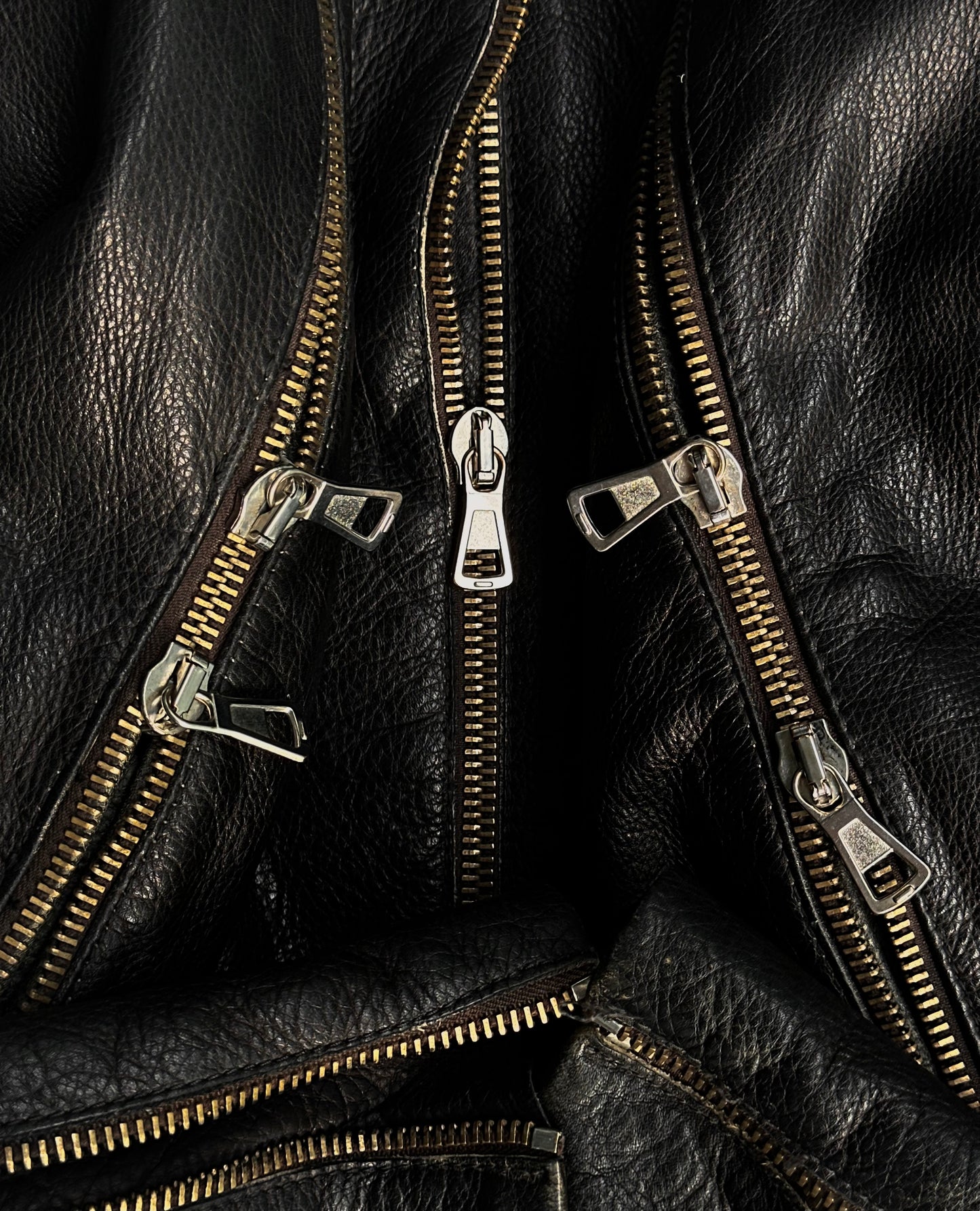 Close-up of metallic zippers on black cowhide leather, inspired by Maison Martin Margiela AW00 Black Cowhide Leather 5-zip Rider Jacket, showcasing shiny teeth and textured grain in an avant-garde arrangement.