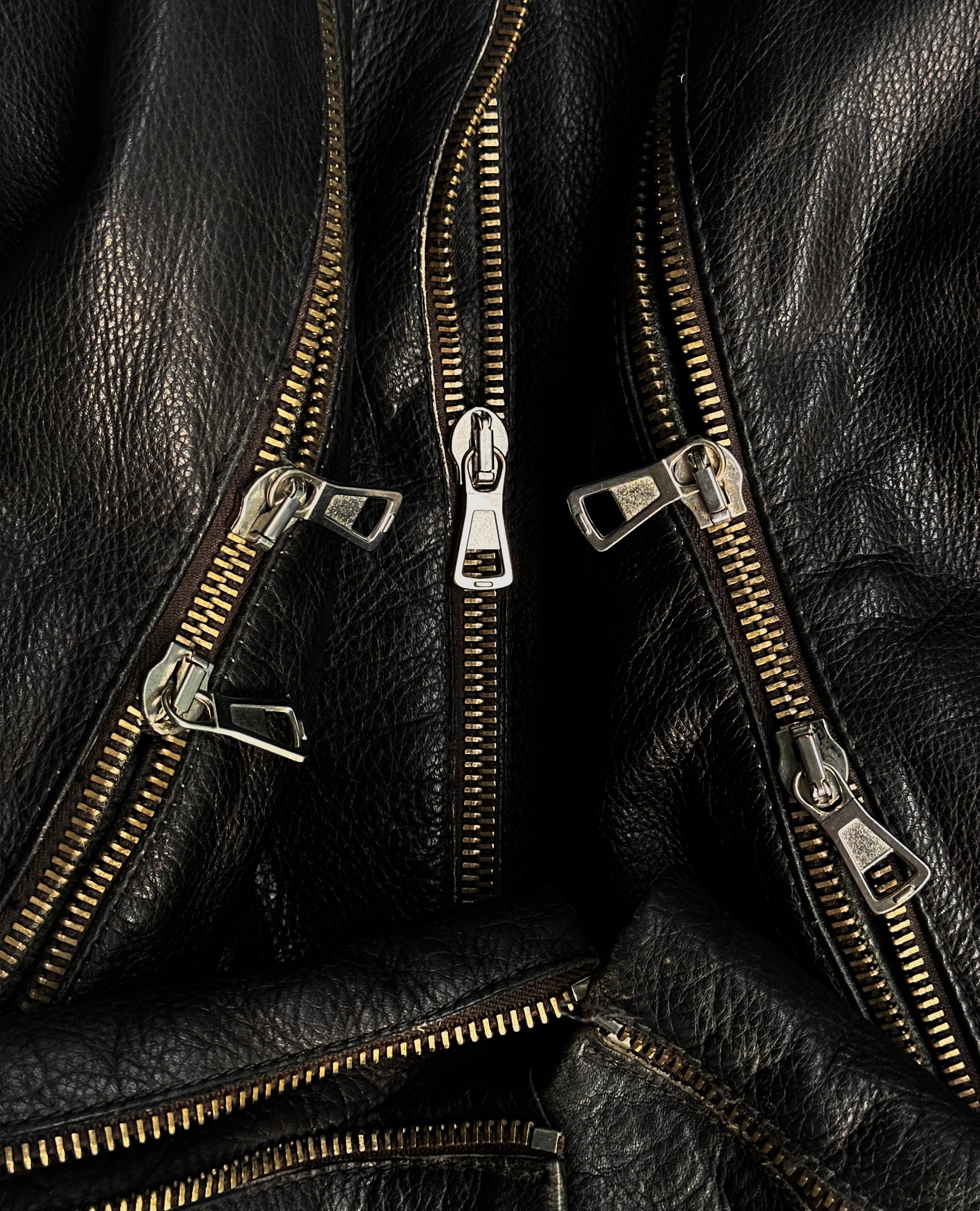 Close-up of metallic zippers on black cowhide leather, inspired by Maison Martin Margiela AW00 Black Cowhide Leather 5-zip Rider Jacket, showcasing shiny teeth and textured grain in an avant-garde arrangement.