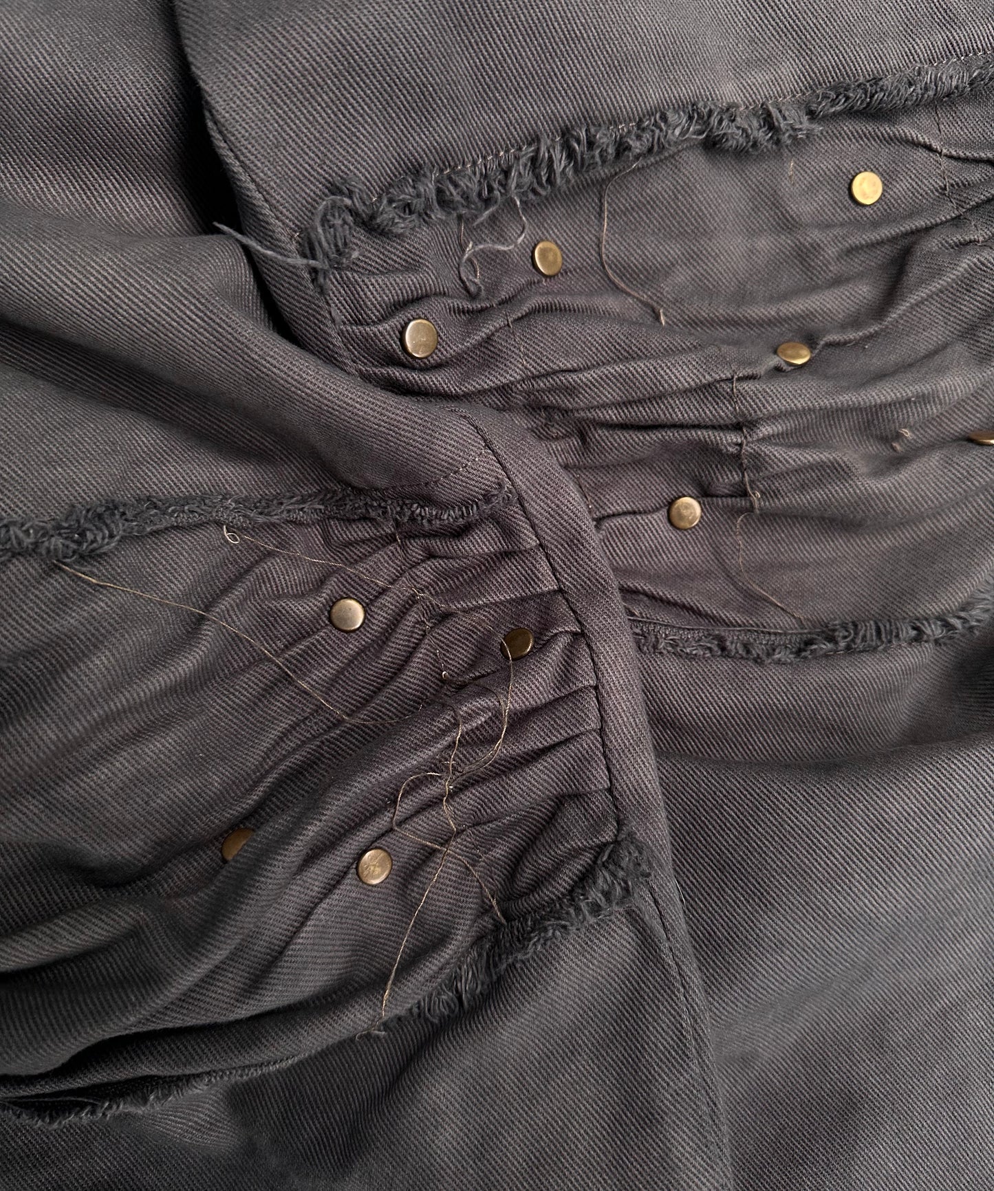 A close-up of ANDREW MACKENZIE Early 00s Stud Darted Knee Denim shows gray fabric with visible seams, loose threads, and rows of small brass studs. The stitching is uneven and the material appears soft with a slightly worn texture.