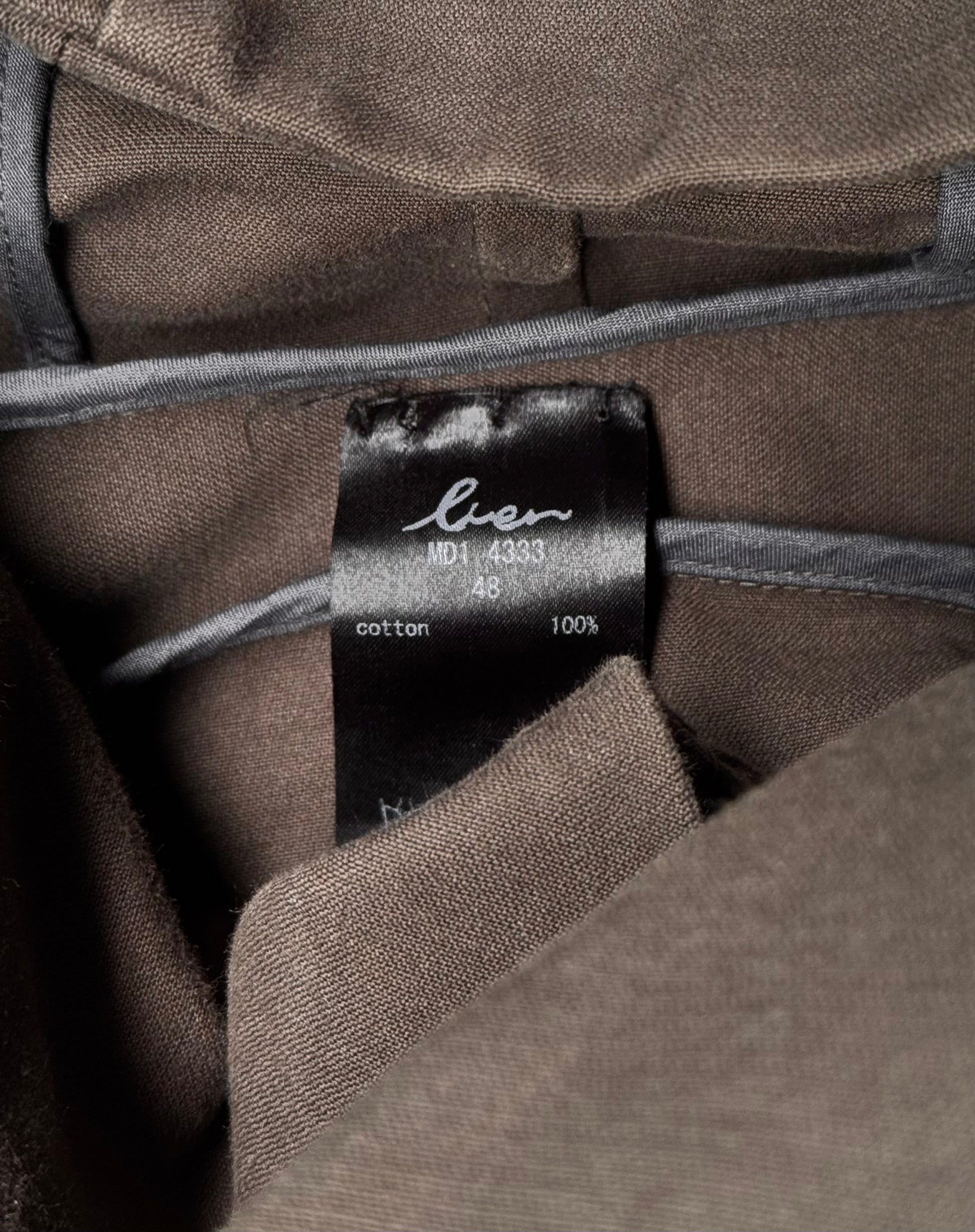 A close-up of a Moleskin Asymmetrical Hooded Mod Coat by lien showcases a neatly folded clothing label. It reads 100% cotton and 48 in white text, with the word bio above in cursive, reminiscent of Japanese avant-garde fashions attention to detail.