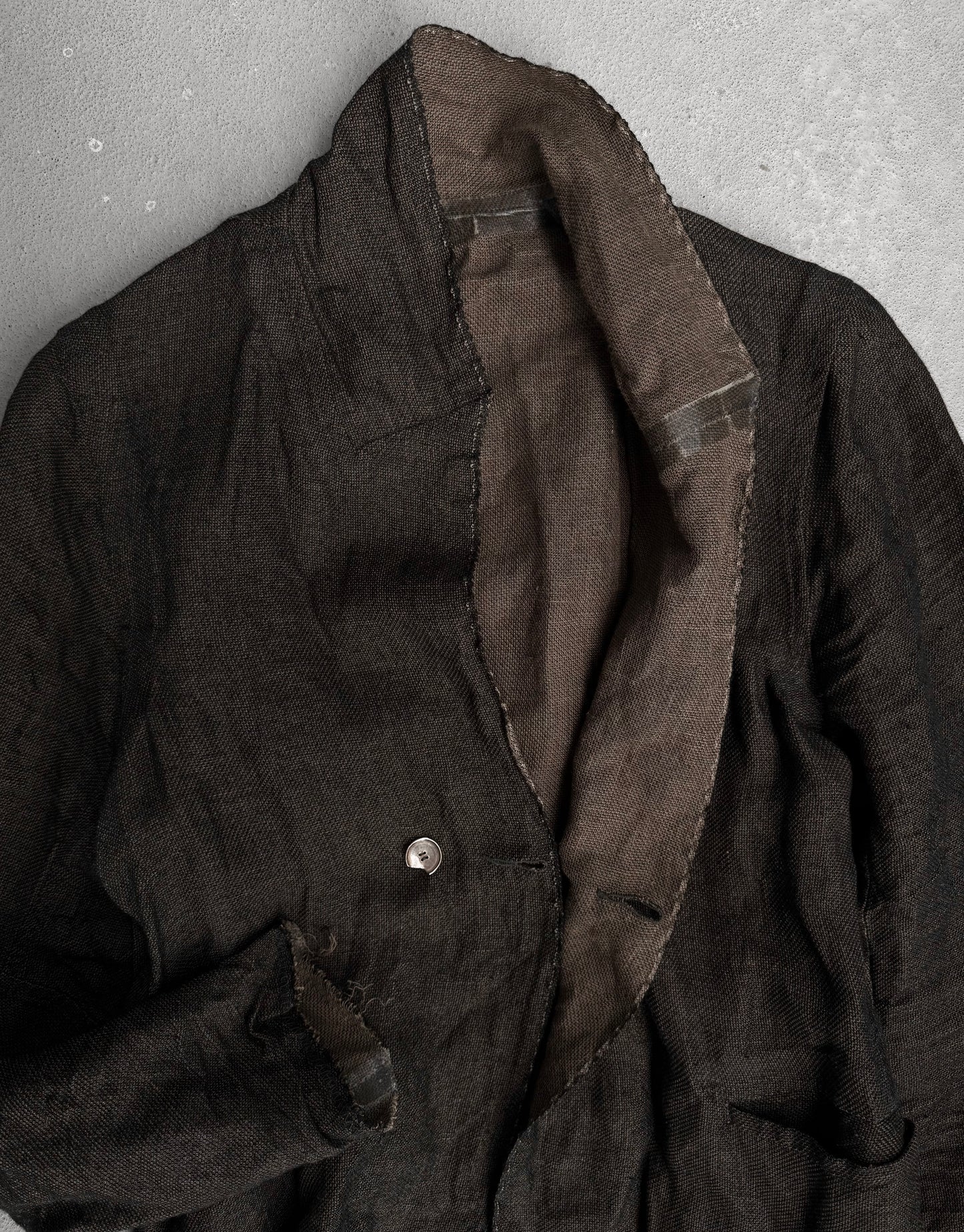 The TaichiMurakami “Coin” Taped Seam Collarless Work Blazer features dark textured Japanese linen, a single visible button, and contrasting brown lining. Its slightly worn look is inspired by industrial minimalism.