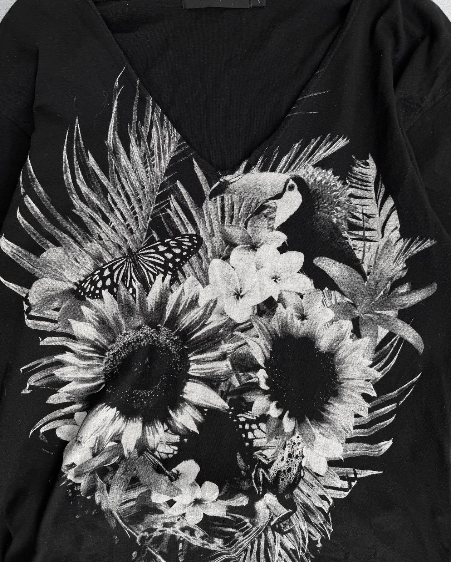 The CIVARIZE Early 00s ‘Floral Skull’ Graphic Long Curve T-shirt features a black and white design with sunflowers, tropical leaves, a toucan, butterfly, and frog in detailed artistry. It channels an edgy Visual Kei-inspired alternative fashion vibe characteristic of the CIVARIZE brand.