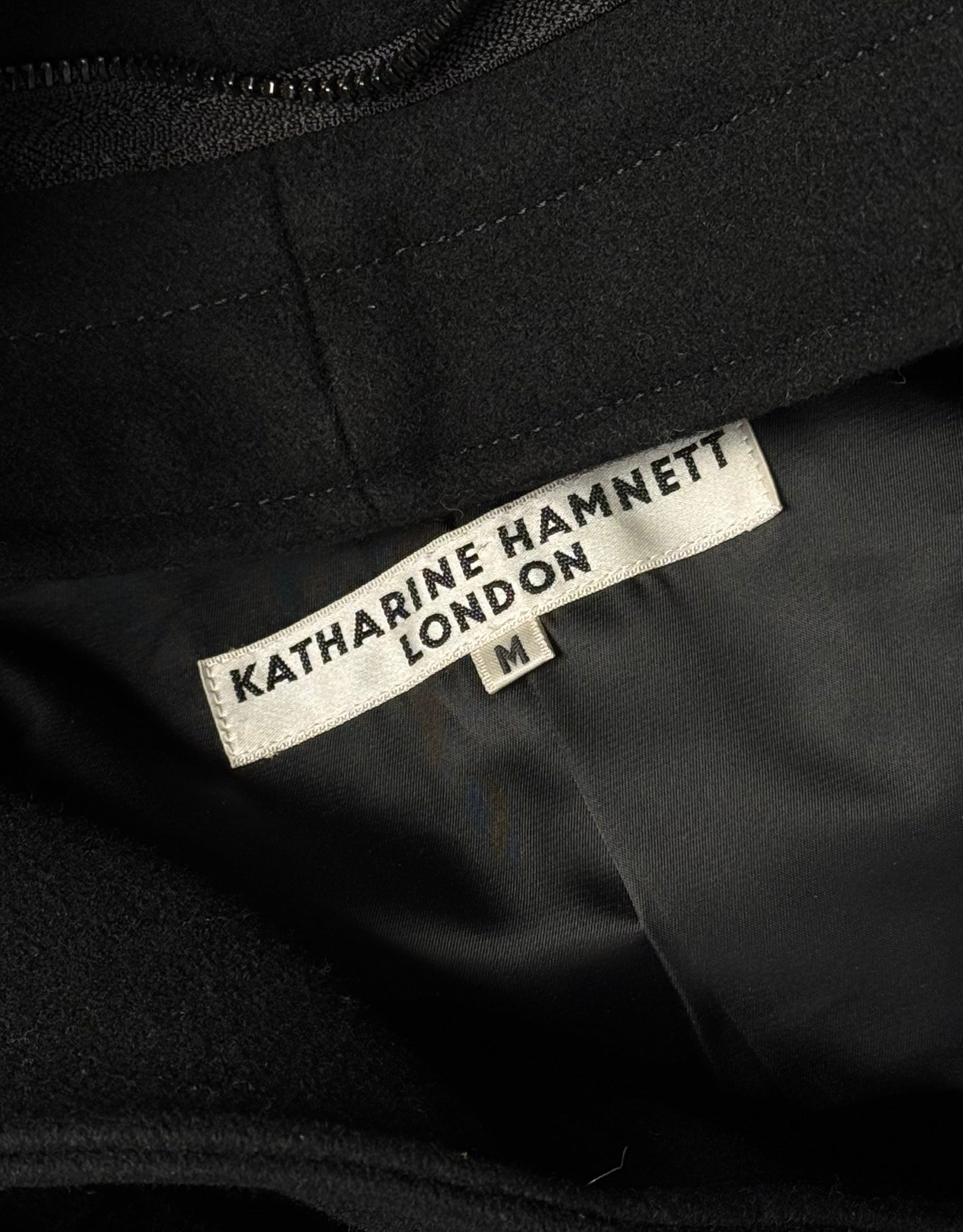 A close-up of a white label sewn onto black fabric, reading “KATHARINE HAMNETT LONDON” in bold, with an “M” size tag—detail from the Early 00s Convertible ‘Alien’ Wool Hooded Rider Jacket by Katharine Hamnett.