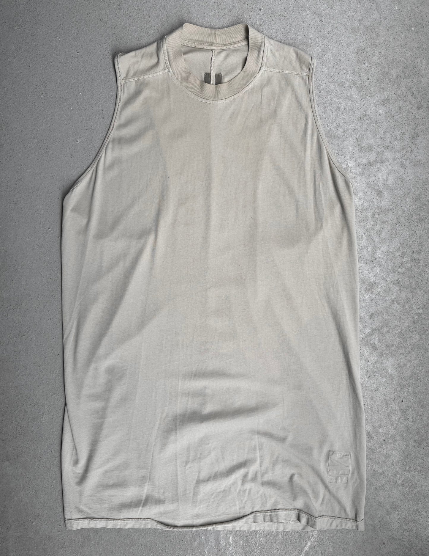 The Rick Owens DRKSHDW AW18 “SISYPHUS” Pearl Racer Tank Top, a beige sleeveless crew-neck style, is laid flat on a gray textured surface. It boasts subtle stitching and a discreet logo near the bottom hem.