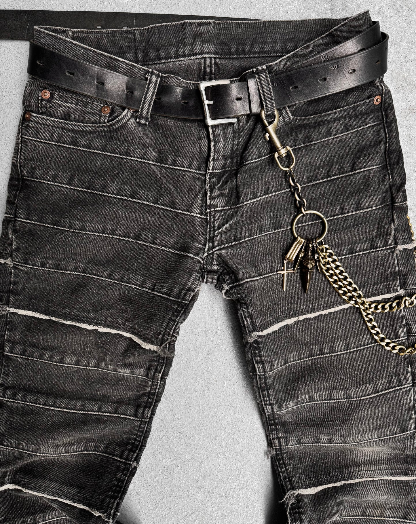 The Hysteric Glamour Early 00s Kinky Grey Flare Hagi Denim features black distressed jeans with a layered, textured design and visible stitching. Complete with a black belt, chain, and keys, it captures punk rock fashion against a neutral gray background.