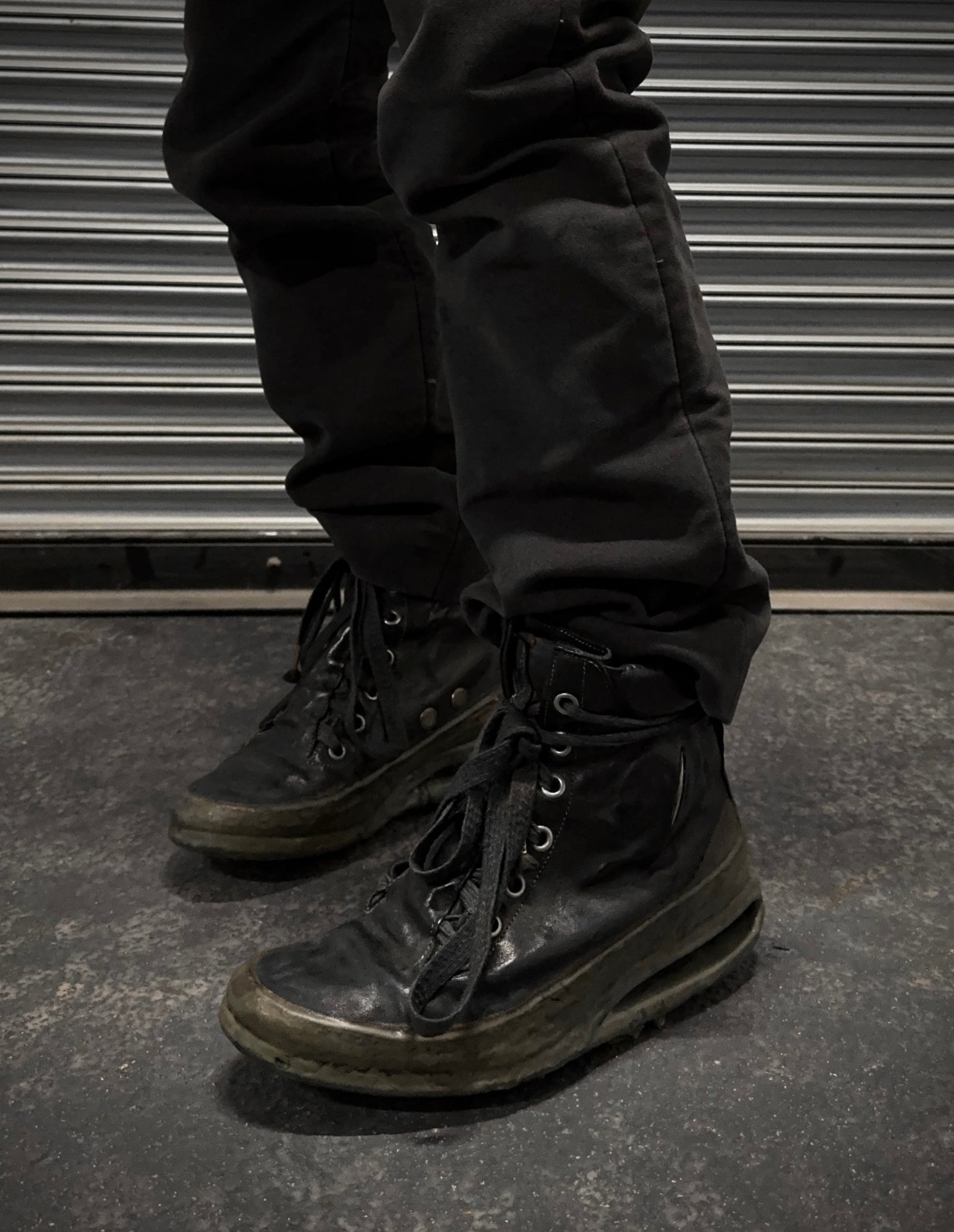A person in black pants and DRIEW Carol Christian Poell ‘AM/2685P ROOMS-PTC/12’ object-dyed kangaroo leather titanium prosthetic U-sole drip sneakers, size 6 with light wear and untied laces, stands on a dark concrete floor by a ribbed metallic shutter.