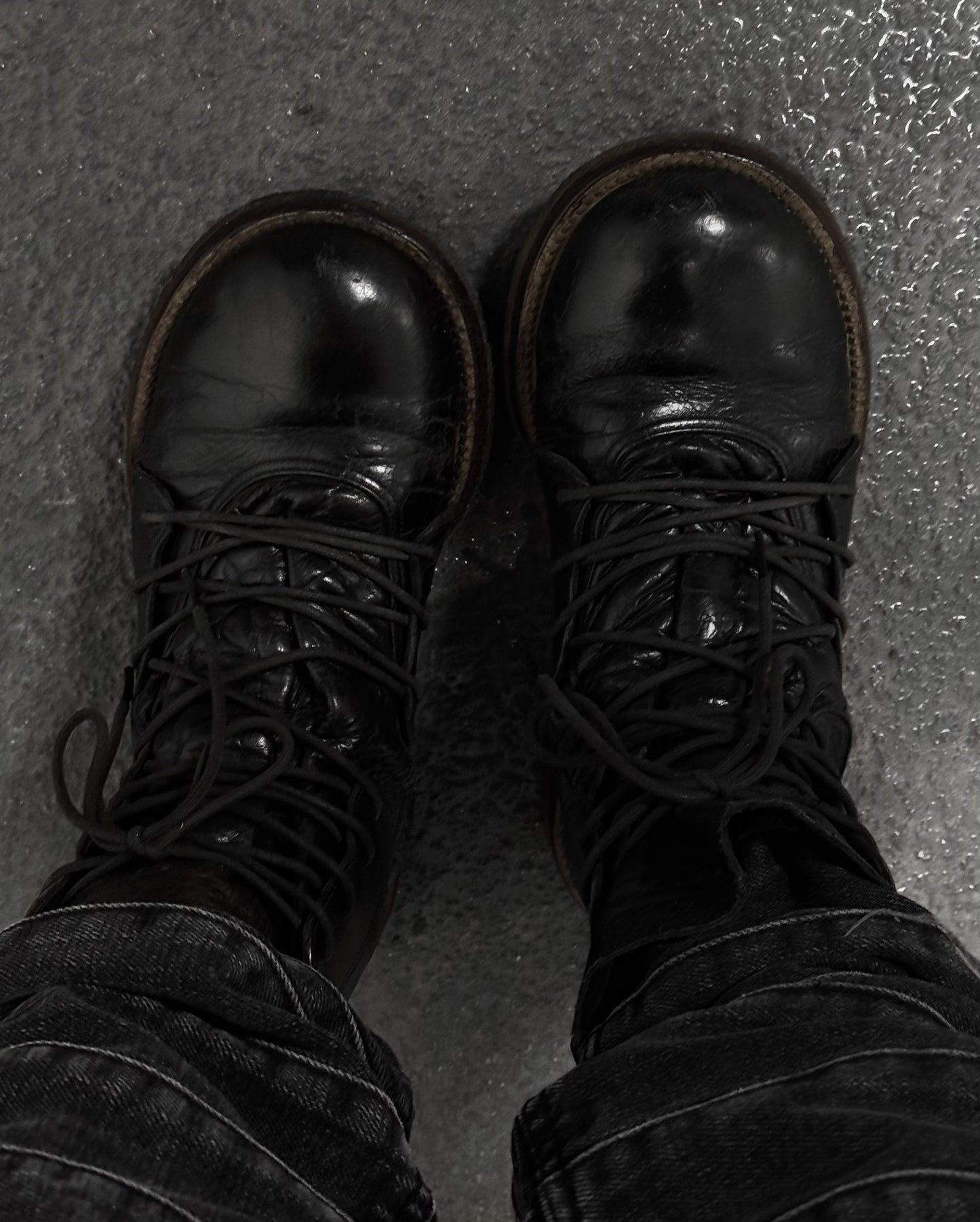 A person in dark jeans stands on a textured gray floor, wearing MiharaYasuhiro Early 00s Multi-laced Crepe Sole Boots by Mihara Yasuhiro. The black leather boots with thick laces showcase avant-garde fashion, featuring a worn shine that hints at frequent use and deconstructed elements.