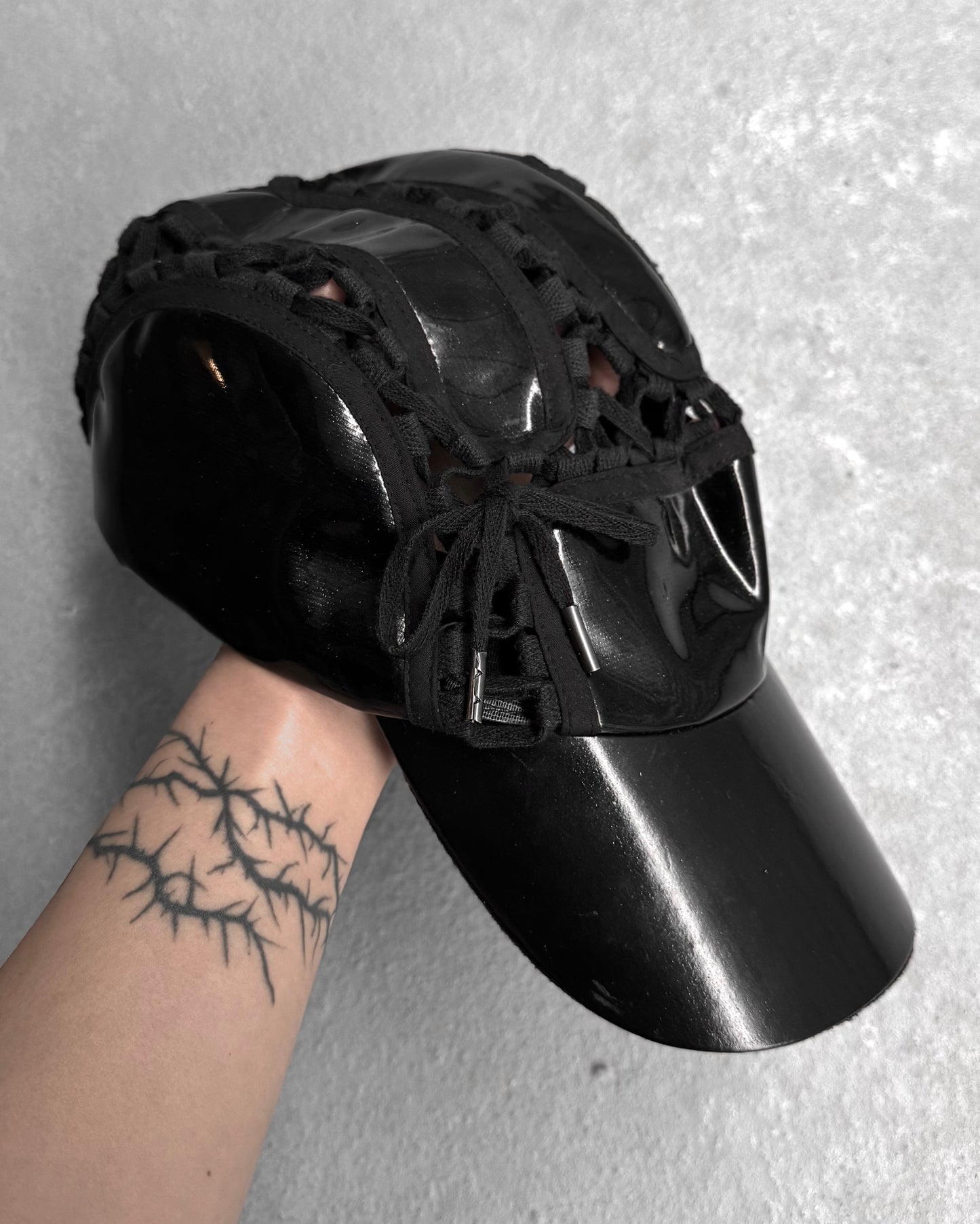 A hand with a thorn-like tattoo holds a black KTZ (Kokon to Zai) Late 00s Lace Through Aviator Cap by London-based brand Konko to Zai, set against a gray textured background.