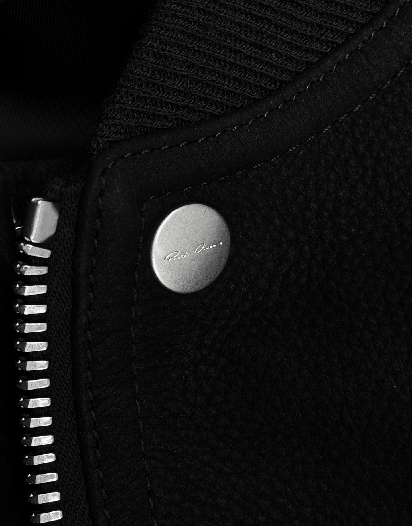 Close-up of the Rick Owens SS24 “LIDO” Nubuck Leather Cropped Flight Bomber Jacket, featuring textured black nubuck leather with a metallic zipper and a round silver snap button engraved with small text.