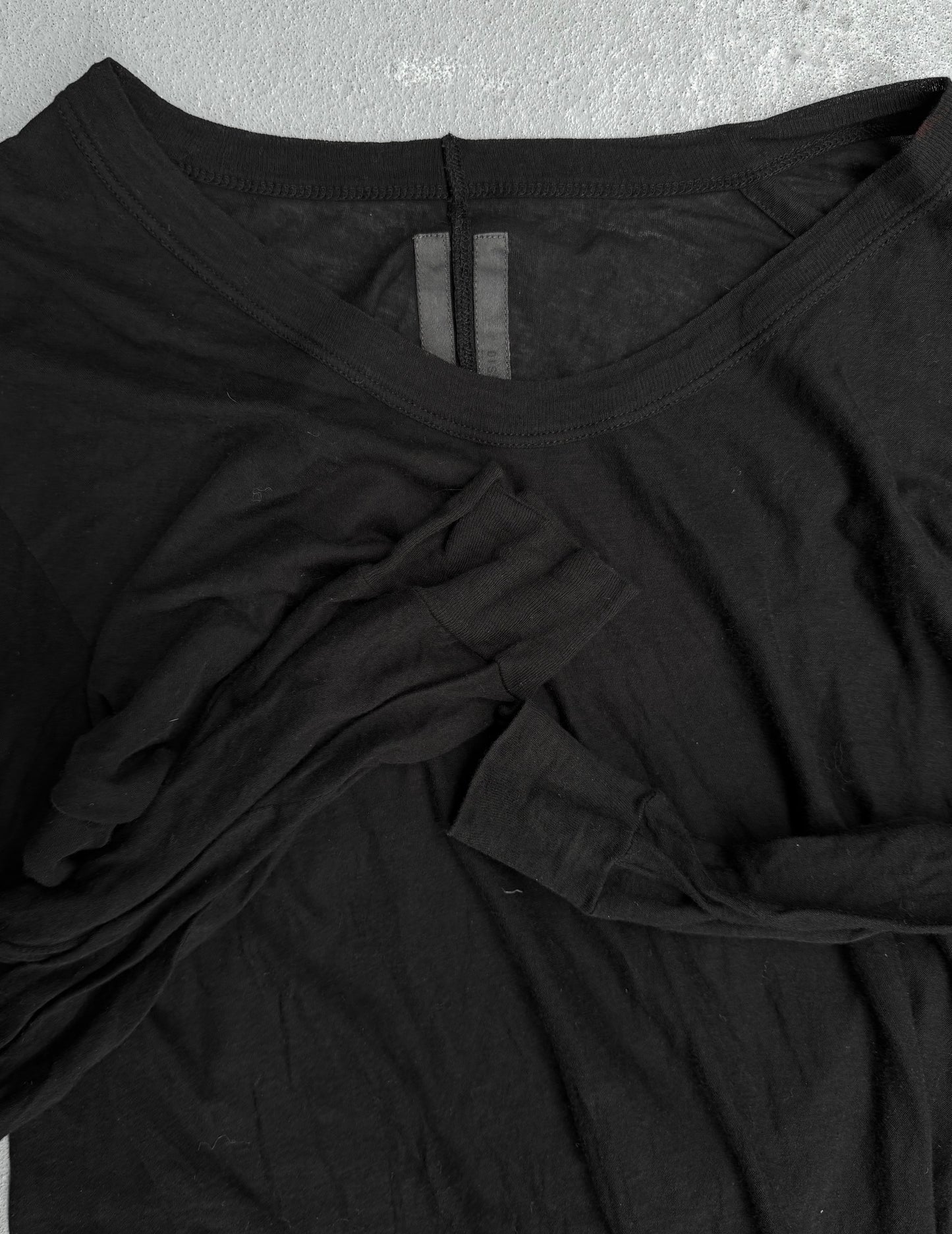 The Rick Owens SS10 “RELEASE” Black Sheer Long-sleeves is laid flat on a gray surface, its long sleeves casually folded across the front, showcasing the brands signature contemporary style.