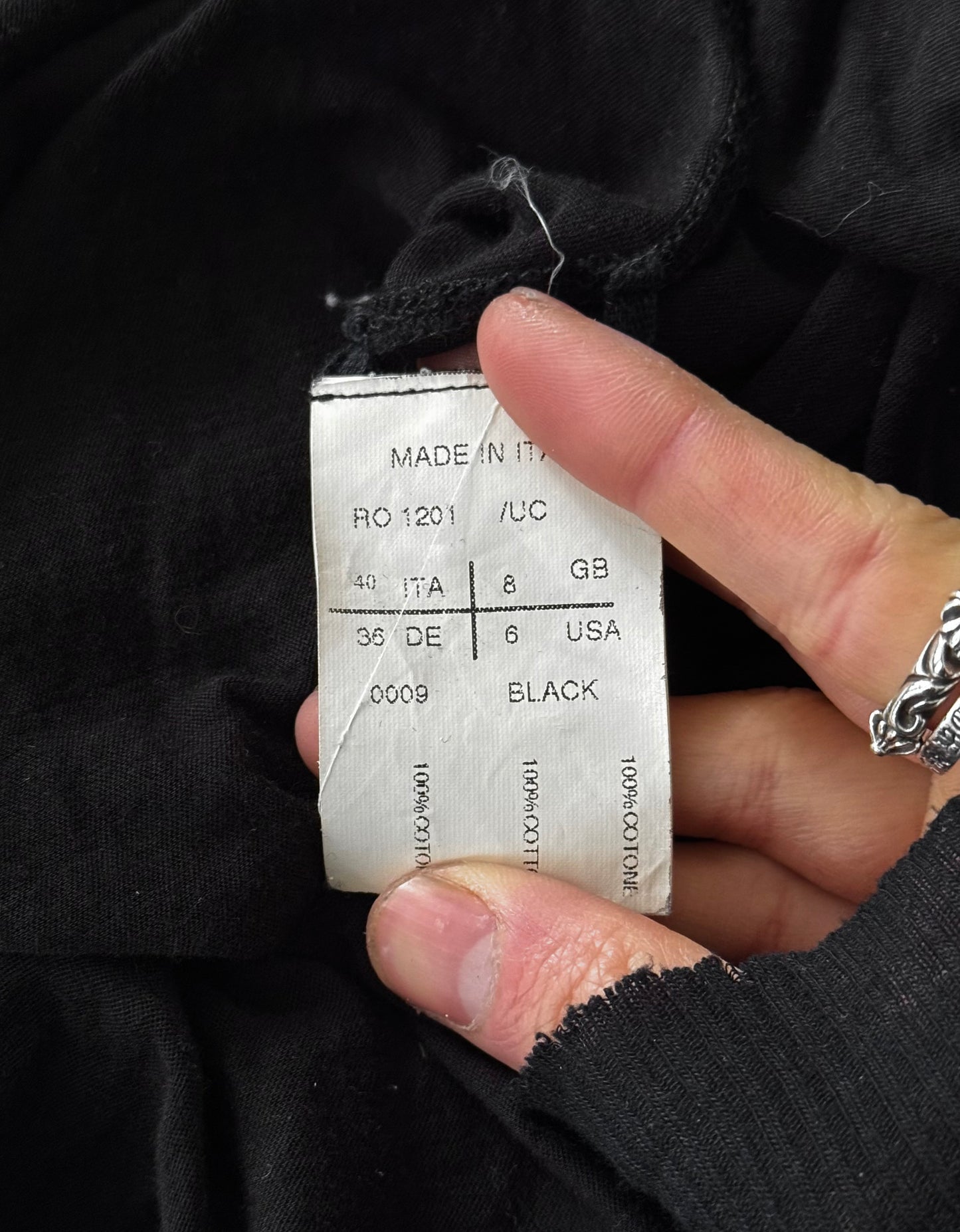 A hand holds the Rick Owens SS10 “RELEASE” Black Sheer Long-sleeves, revealing a white tag with sizing for Italy, UK, and USA: BLACK, 100% cotton, MADE IN ITALY—showcasing Rick Owens signature contemporary craftsmanship.