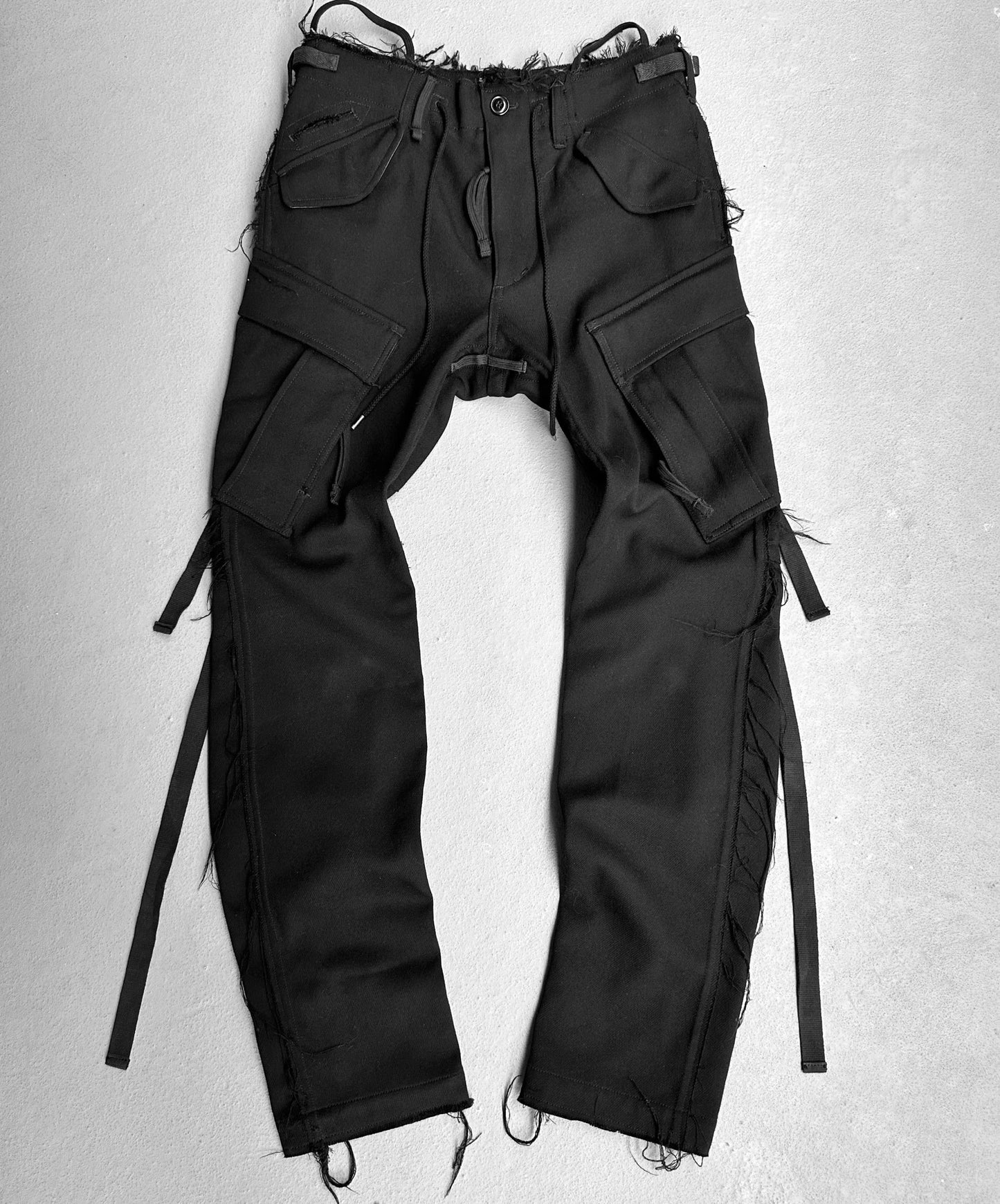 On a flat surface lie the TakahiroMiyashita TheSoloist. AW15 Calf-trimmed Wool ‘Bondage’ Straps Cargo Pants, featuring a distressed waist, multiple large pockets, and side straps that echo Takahiro Miyashitas rugged style and avant-garde fashion.