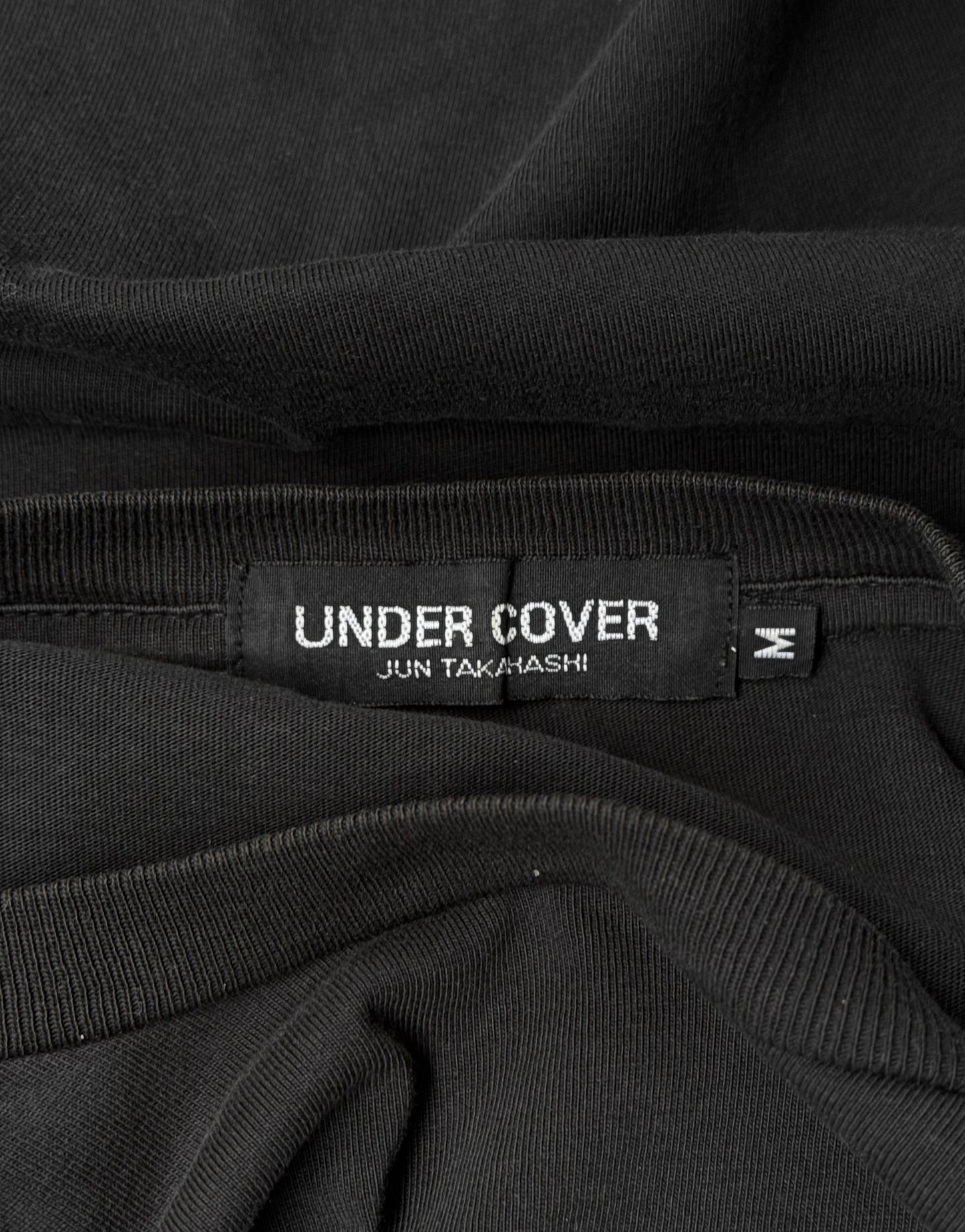 A close-up of a black UNDERCOVER label reading JUN TAKAHASHI stitched onto an Undercover SS16 ‘Gilapple’ Graphic Printed T-shirt, with size 2 shown on a small tag, highlighting iconic streetwear design.