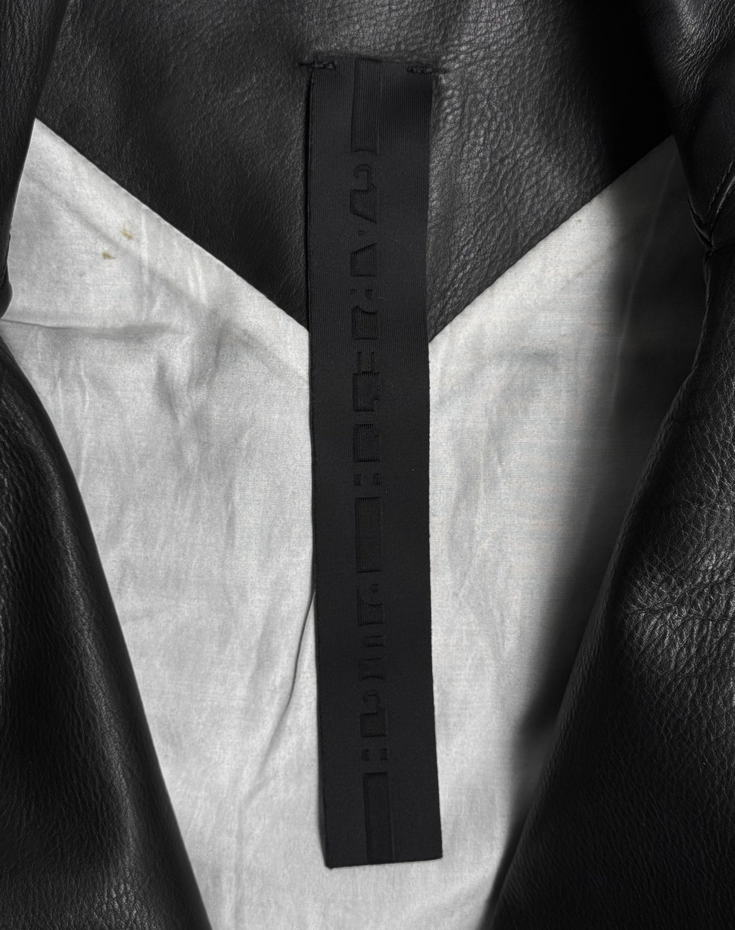 Close-up of the inside collar of a Gareth Pugh AW09 Detachable Sleeves Horse Leather Rider’s Jacket, showing a black tag with embossed, unreadable characters and a light gray inner lining.