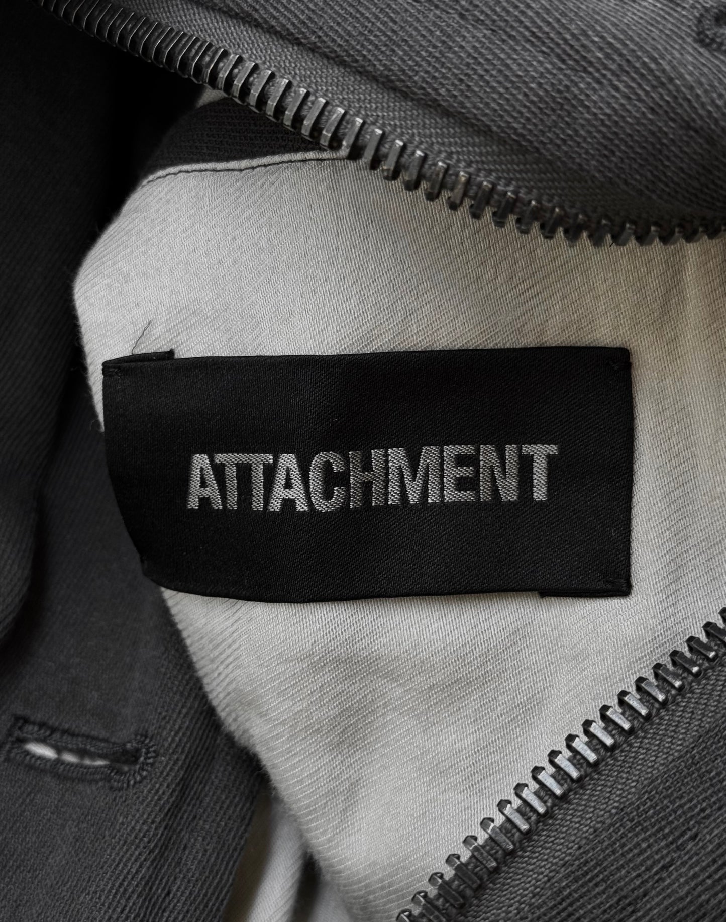 A close-up of the ATTACHMENT by Kazuyuki Kumagai label, sewn onto a light fabric Raglan Doctor Trench Coat, partially framed by an open gray zipper.