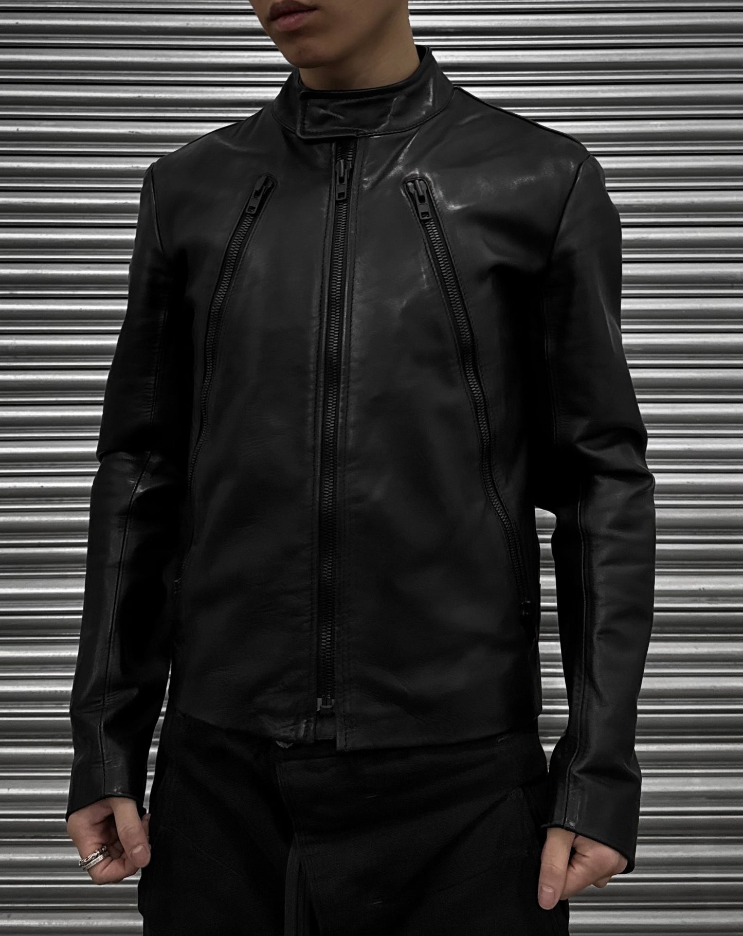 A person models a Maison Martin Margiela AW10-11 Kokone Black Horse Leather 5-zip Jacket (size 44) with vertical zippers, standing before a metallic corrugated shutter. The photo is cropped above the mouth and below the waist.