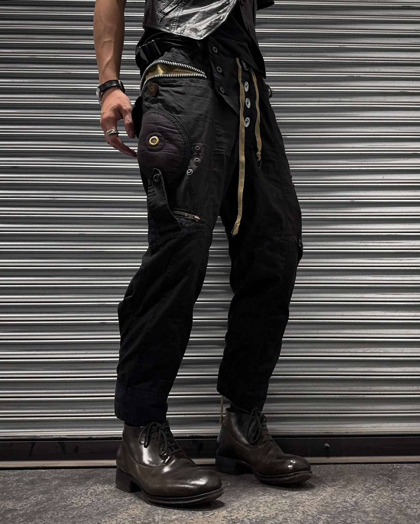 A person stands before a metal shutter wearing L.G.B Early 00s Pilot Cargo Capris (US 32 Waist, used), featuring repair patches, button details, and a belt with metal rings. They pair them with dark boots and several rings and bracelets; torso is partially out of frame.