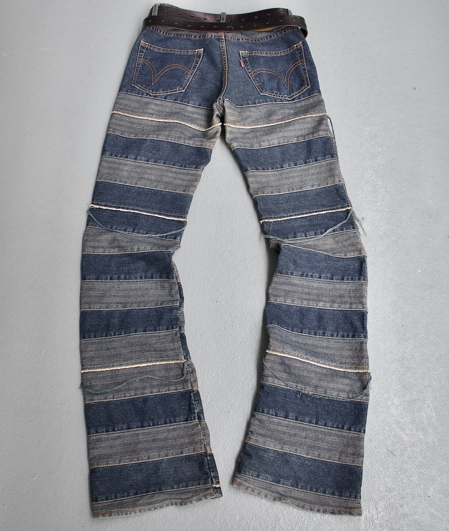 The Hysteric Glamour Early 00s ‘Kinky’ Blue Hagi Flare Denim features alternating dark and light blue striped patches, visible seams, and a brown belt, all embodying punk rock fashion. Displayed on a light gray surface, these jeans reflect the rebellious spirit of Hysteric Glamour.