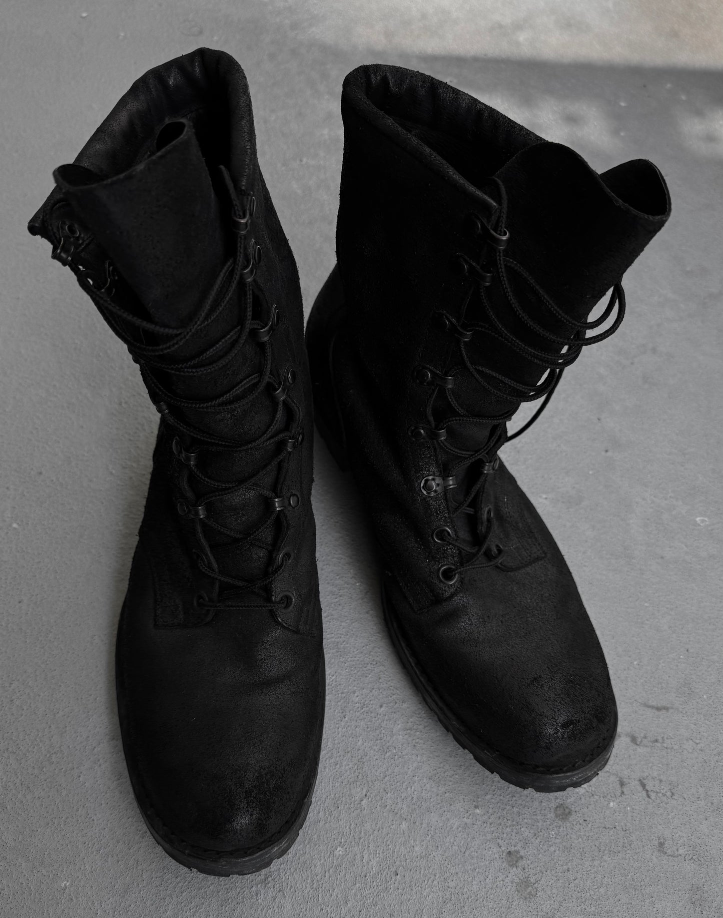 A pair of Attachment by Kazuyuki Kumagai Reversed Cow Leather Lace-up Boots with undone laces, standing on a gray concrete surface.