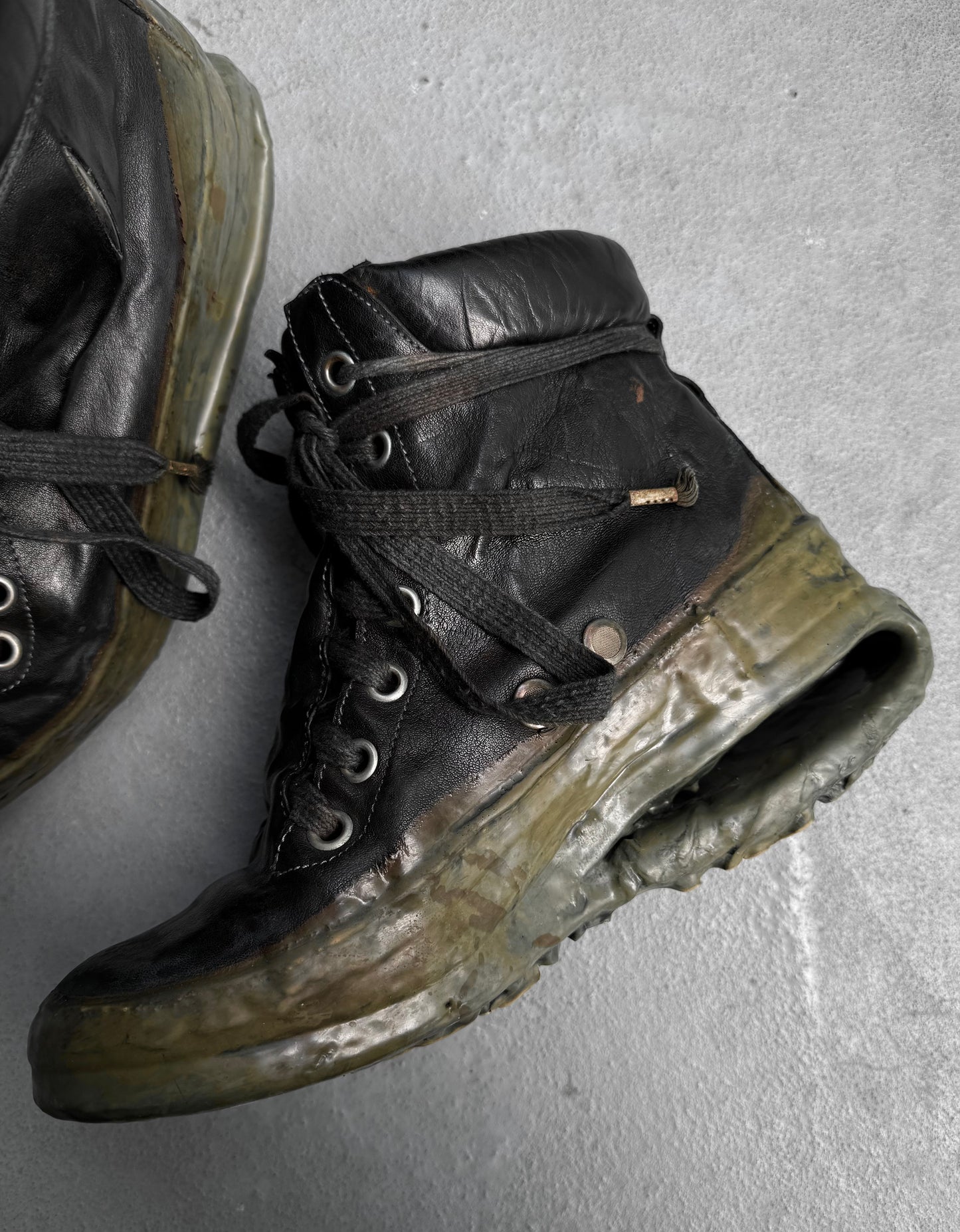 A worn DRIEW Carol Christian Poell ‘AM/2685P ROOMS-PTC/12’ black kangaroo leather sneaker, Size 6, with a cracked, melted titanium U-sole is displayed on a gray surface; part of another similarly damaged sneaker is seen in the corner.