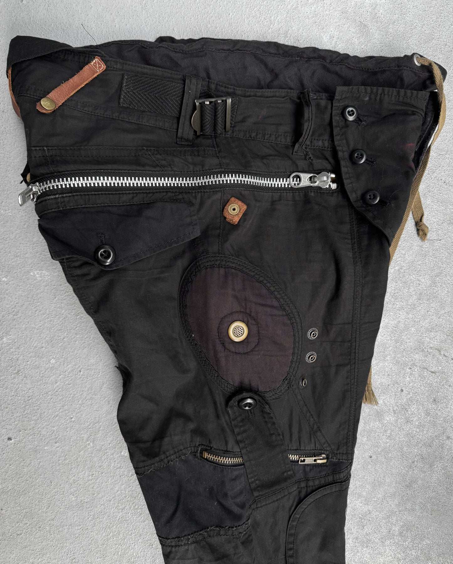 L.G.B Early 00s Pilot Cargo Capris by L.G.B, US 32 Waist, black with multiple zippers, buttons, repair patches, and pockets. Features an asymmetrical design and varied fabric textures. Minor staining visible. Laid flat on a gray surface.