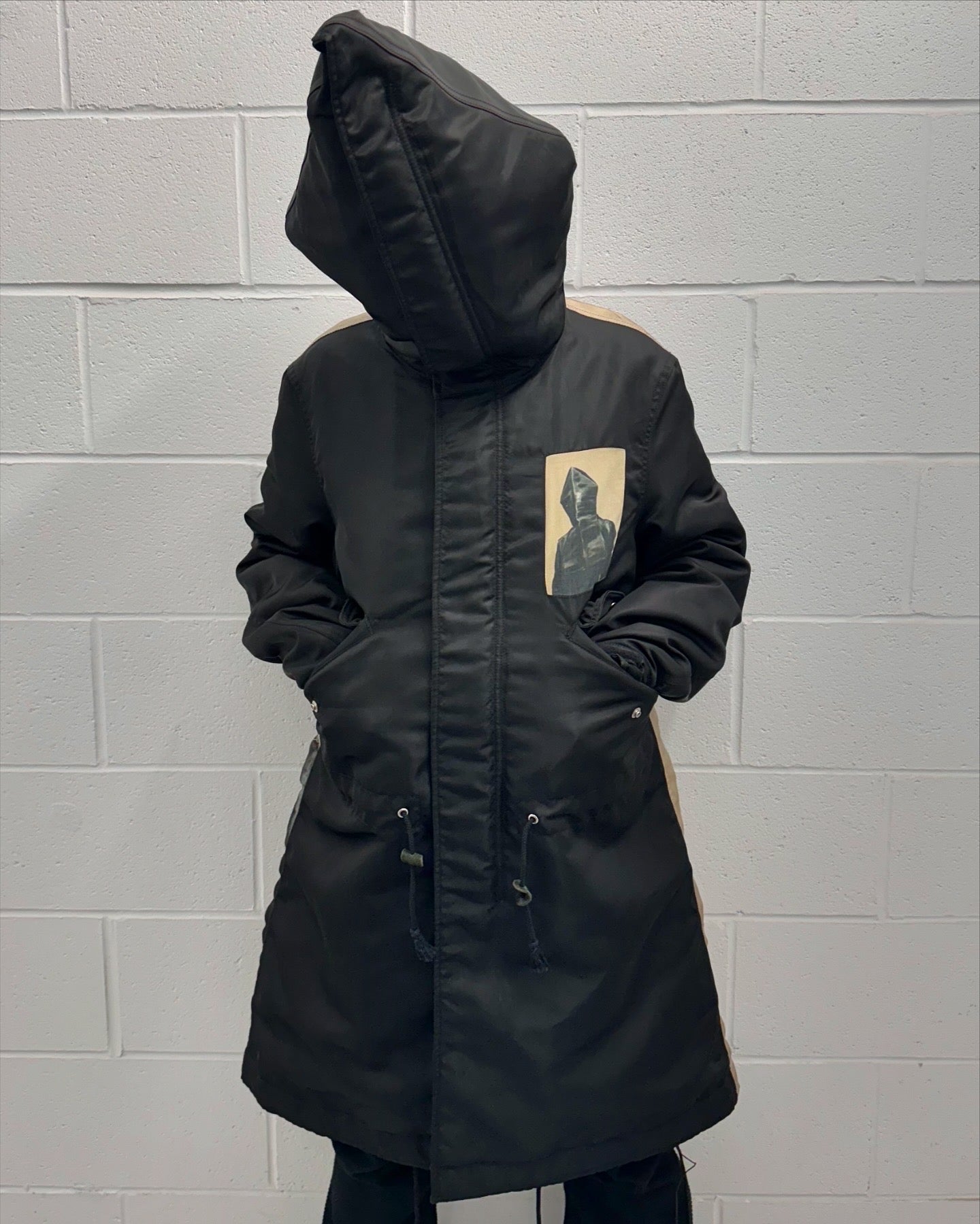 A person in an UNDERCOVER AW16 ‘Borremans’ Full-zip Fishtail Nylon Parka by Undercover, featuring a hood and beige patch with a hooded figure on the chest, stands against a white brick wall with their face concealed by the hood.