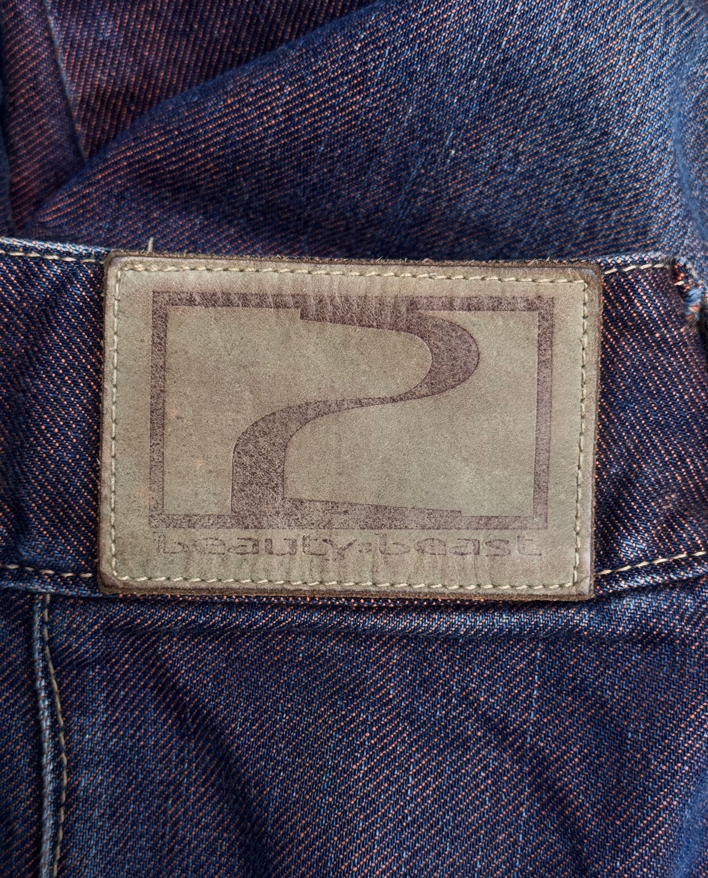 Close-up of a brown leather patch on Beauty:Beast Late 90s Over-dyed Articulate Bootcut Denim, showcasing avant-garde Tokyo fashion. The stylized logo resembles a curved road with partially visible text beneath it, and meticulous stitching along the edges reminiscent of Beauty:Beast craftsmanship.