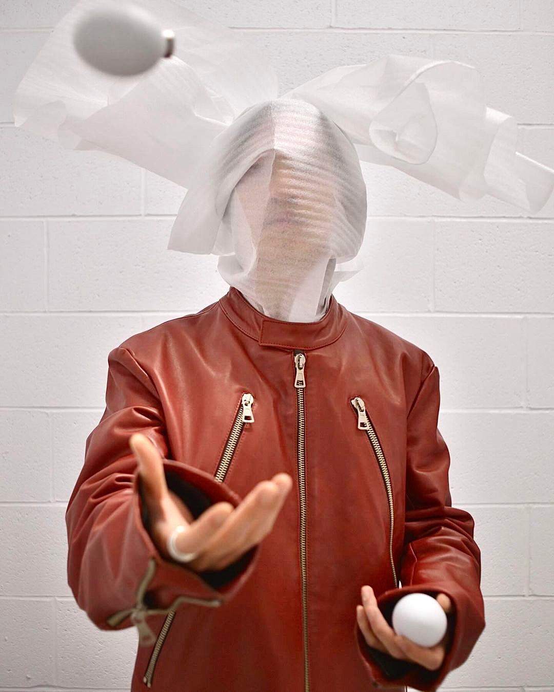 A person in a Maison Martin Margiela AW00 Red Cowhide Leather 5-zip Rider Jacket with their face veiled in white fabric tosses two white eggs against a white brick wall, evoking the brand’s signature avant-garde style.