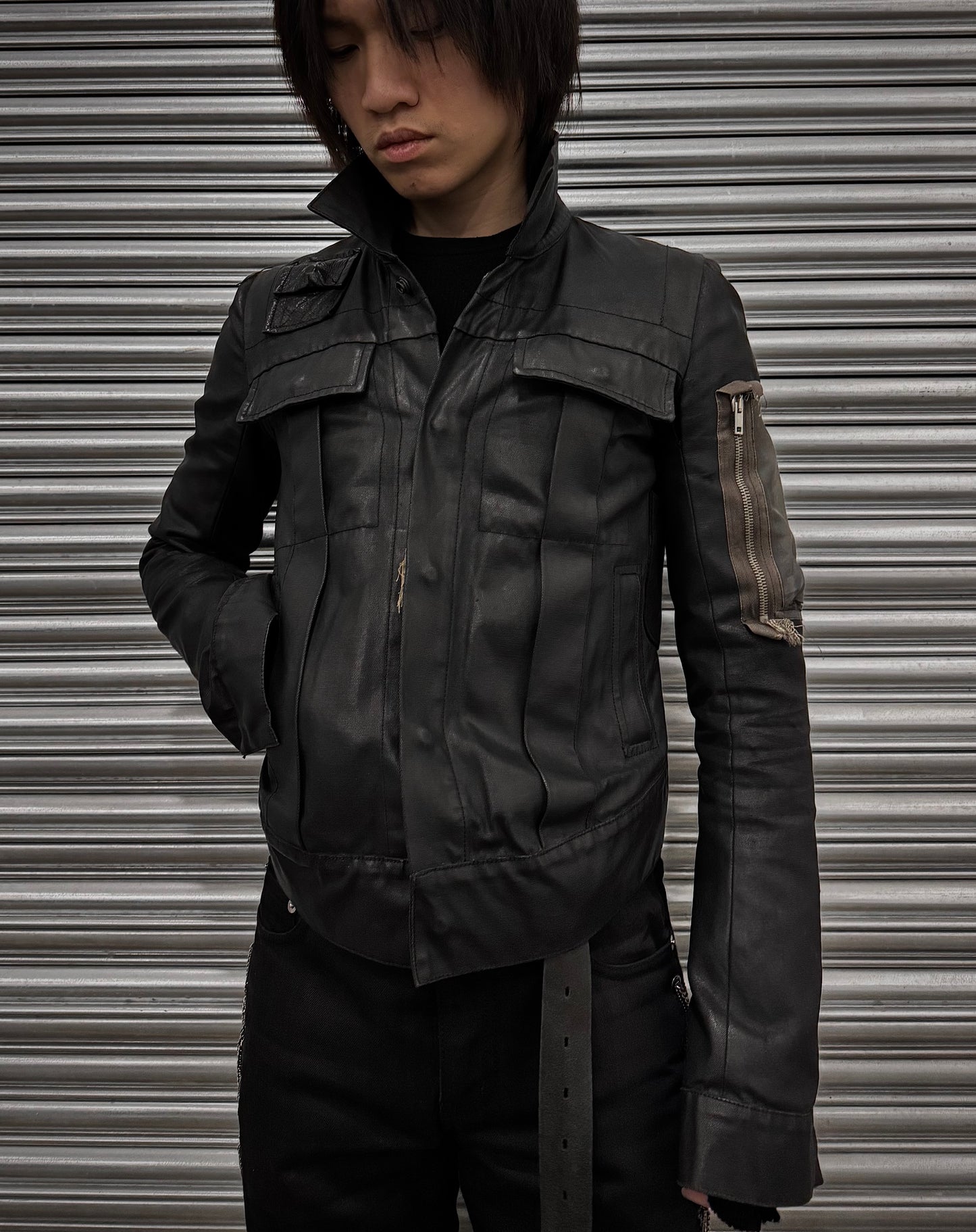 In front of a closed metal roller door, a person with medium-length dark hair and hands in pockets wears a Rick Owens SS03 SUCKERBALL Resinated Denim MA-1 Trucker Jacket, featuring a sleeve zipper and exuding the brands avant-garde style.