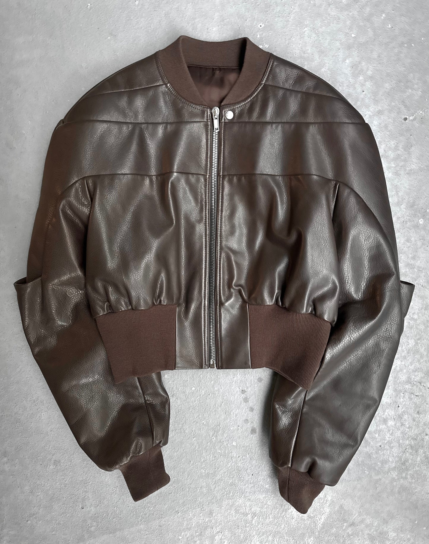 The Rick Owens AW23 LUXOR Girdered Leather Bomber Jacket, in cropped brown calfskin with a front zipper and ribbed collar, cuffs, and waistband, is shown laid flat on a gray surface.