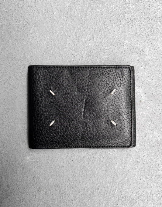 The Maison Martin Margiela SS14 Tumbled Leather Bifold Wallet features black textured leather and four white stitches in a rectangular pattern, reflecting the brand’s avant-garde style, set on a light gray concrete background.