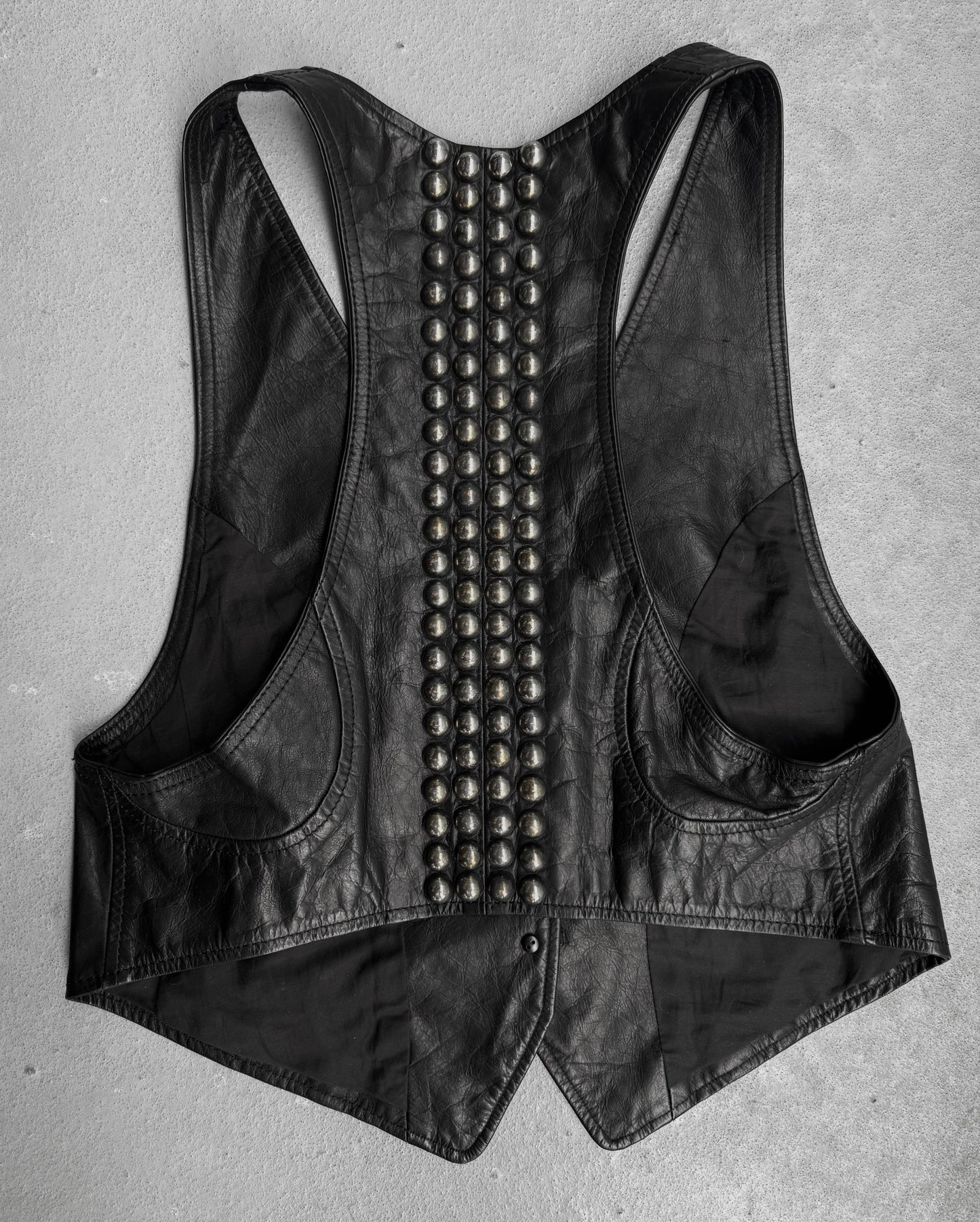 The L.G.B Early 00s Calfskin Leather Studded Mini-vest by L.G.B is a black racerback vest with two vertical rows of large metallic studs down the back, inspired by Japanese fashion and shown on a light gray surface.