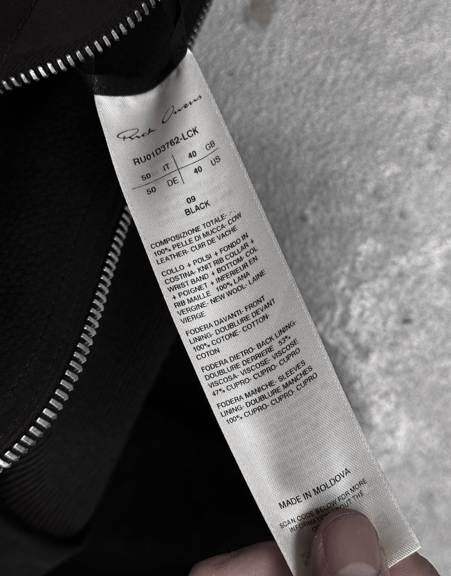 A hand holds a white label with black text showing fabric details, care instructions, size, and MADE IN MOLDOVA on a Rick Owens SS24 “LIDO” Nubuck Leather Cropped Flight Bomber Jacket near a black zipper against a grey background.