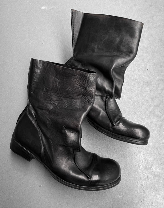 A pair of black Boris Bidjan Saberi x Muñoz Vrandecic AW14-15 ‘BOOT1-F235’ horse leather pull-on boots with an asymmetrical, wrinkled design sits on a light gray surface.