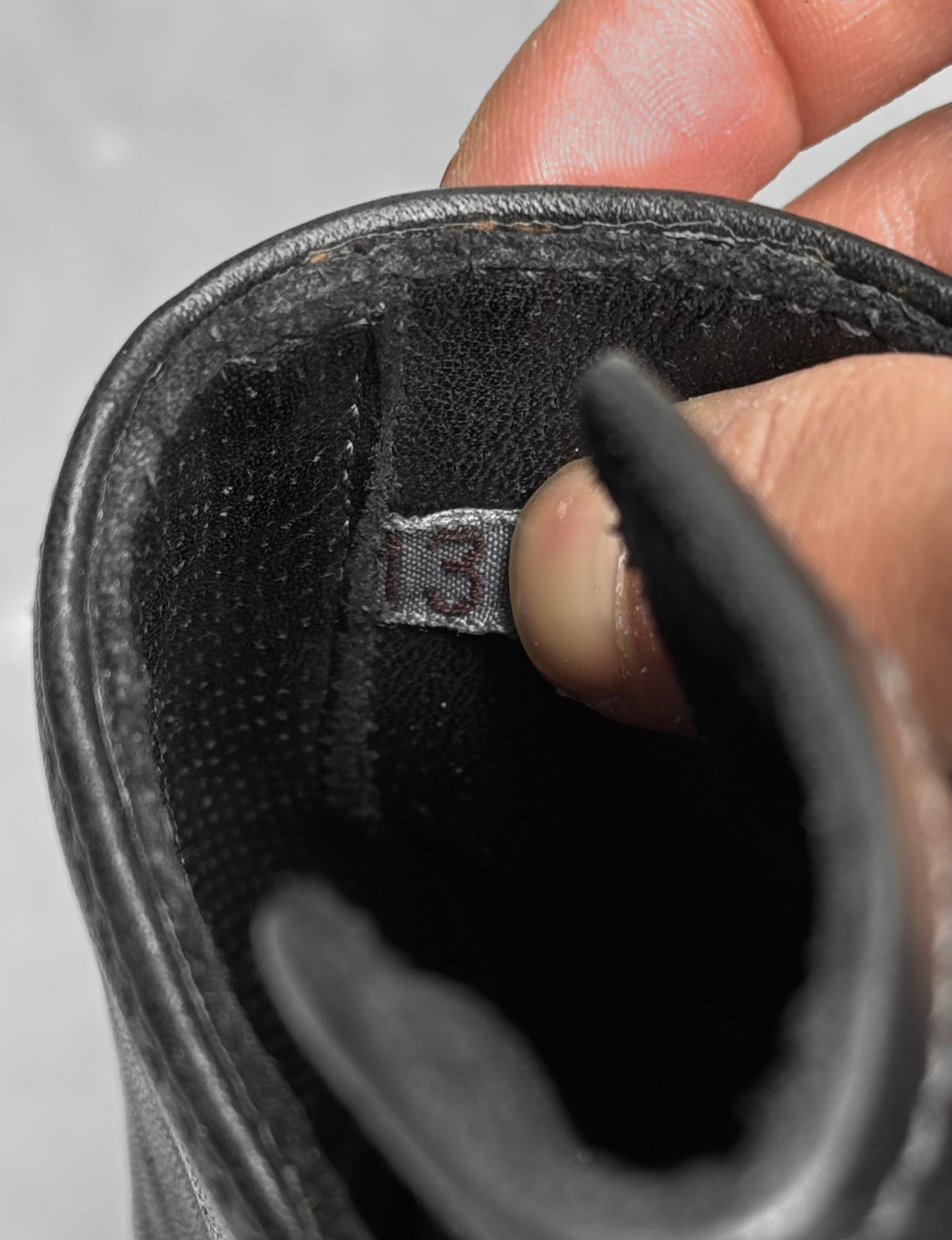 A close-up of a hand holding open the tongue of a new Carol Christian Poell ‘AM/2759SP-IN CORSS-PTC’ Kangaroo Leather Buckle Drip Boot, revealing a fabric tag with the number 13.