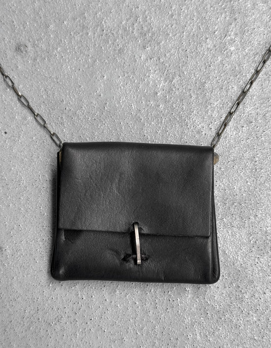 The M.A+ by Maurizio Amadei ‘A-B7201’ Calfskin Leather Mini Accordion Pouch Necklace, OneSize in black with a metal chain strap and bar clasp, is showcased on a textured gray concrete surface.