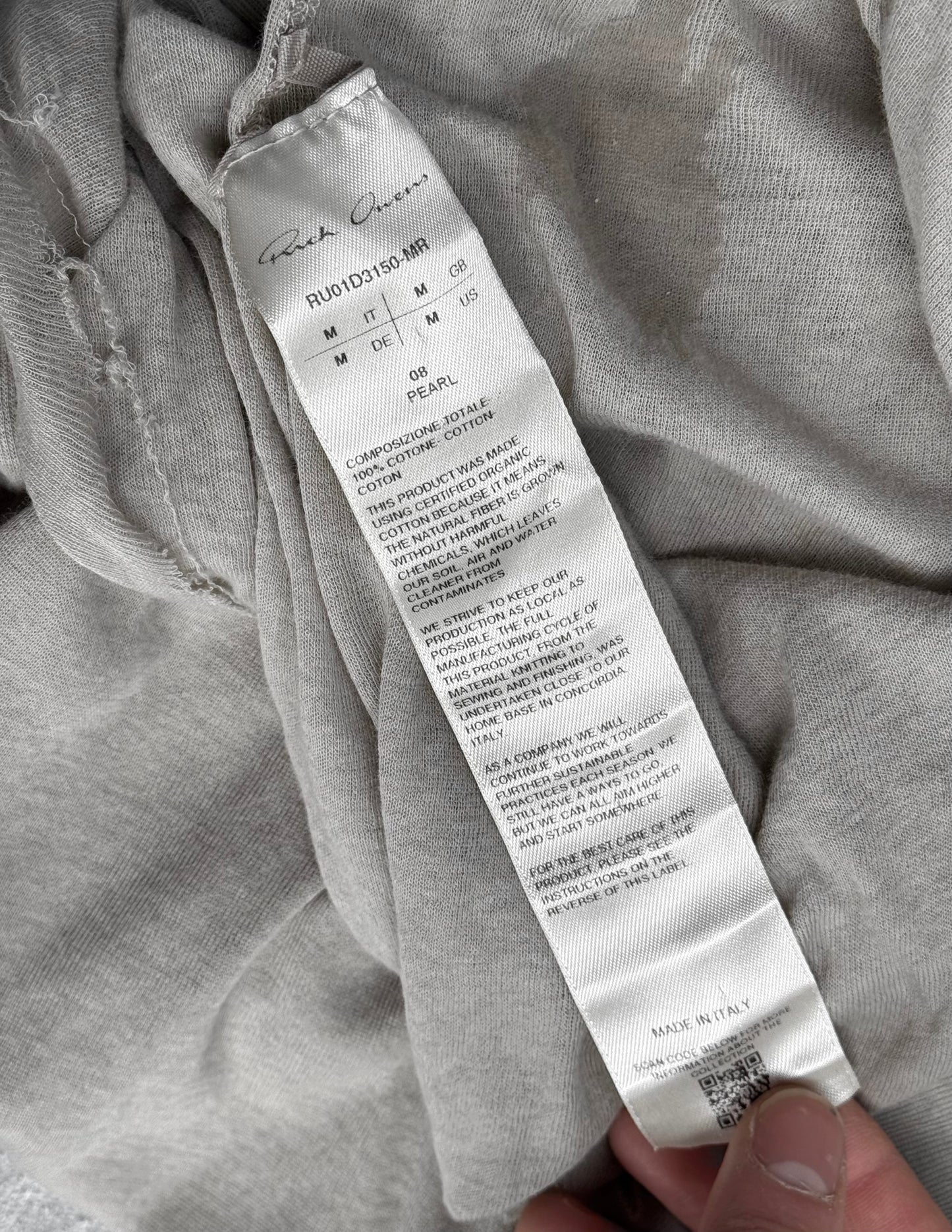 A close-up of a care label on the Rick Owens SS24 “LIDO” Pearl Sheer D-Flap Tank Top, held between fingers. The label lists washing instructions and materials—showcasing the quality typical of Rick Owens innovative designs.