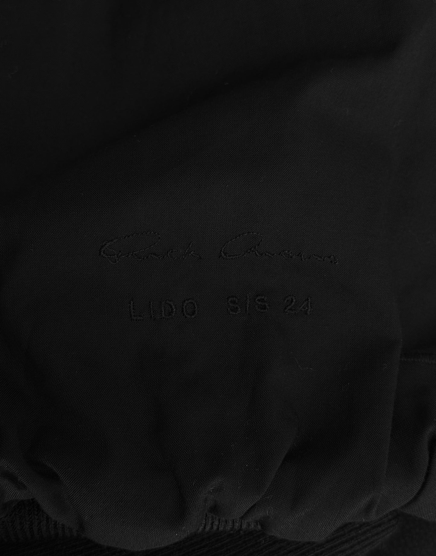 A close-up of black nubuck leather with subtle embossed LIDO SS24 text and a faint cursive signature, capturing the avant-garde style of the Rick Owens SS24 “LIDO” Cropped Flight Bomber Jacket.