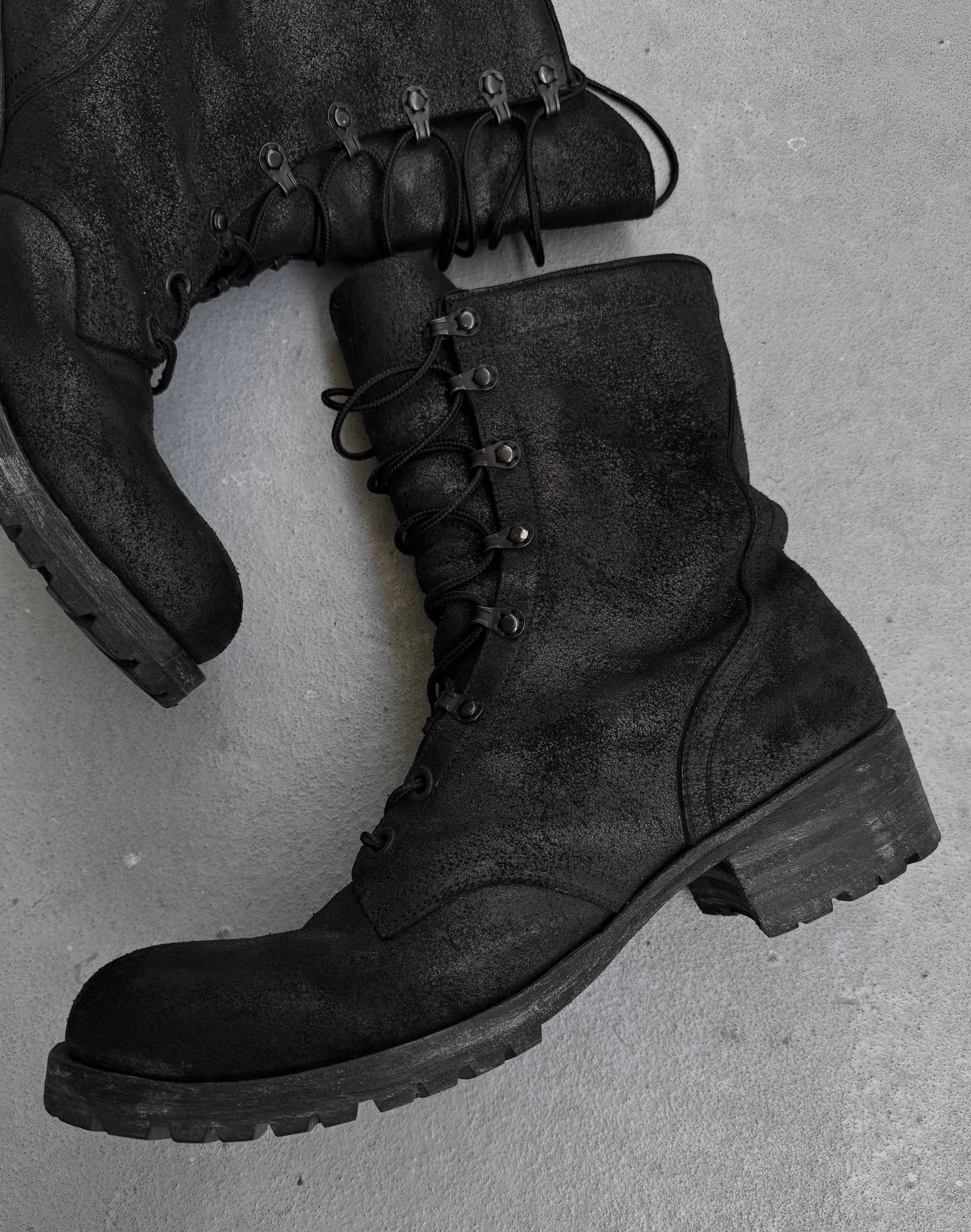 ATTACHMENT by Kazuyuki Kumagai Reversed Cow Leather Lace-up Boots: black combat boots with thick, rugged soles, one upright and one on its side, partially overlapping on a light grey surface. Perfect for menswear enthusiasts.